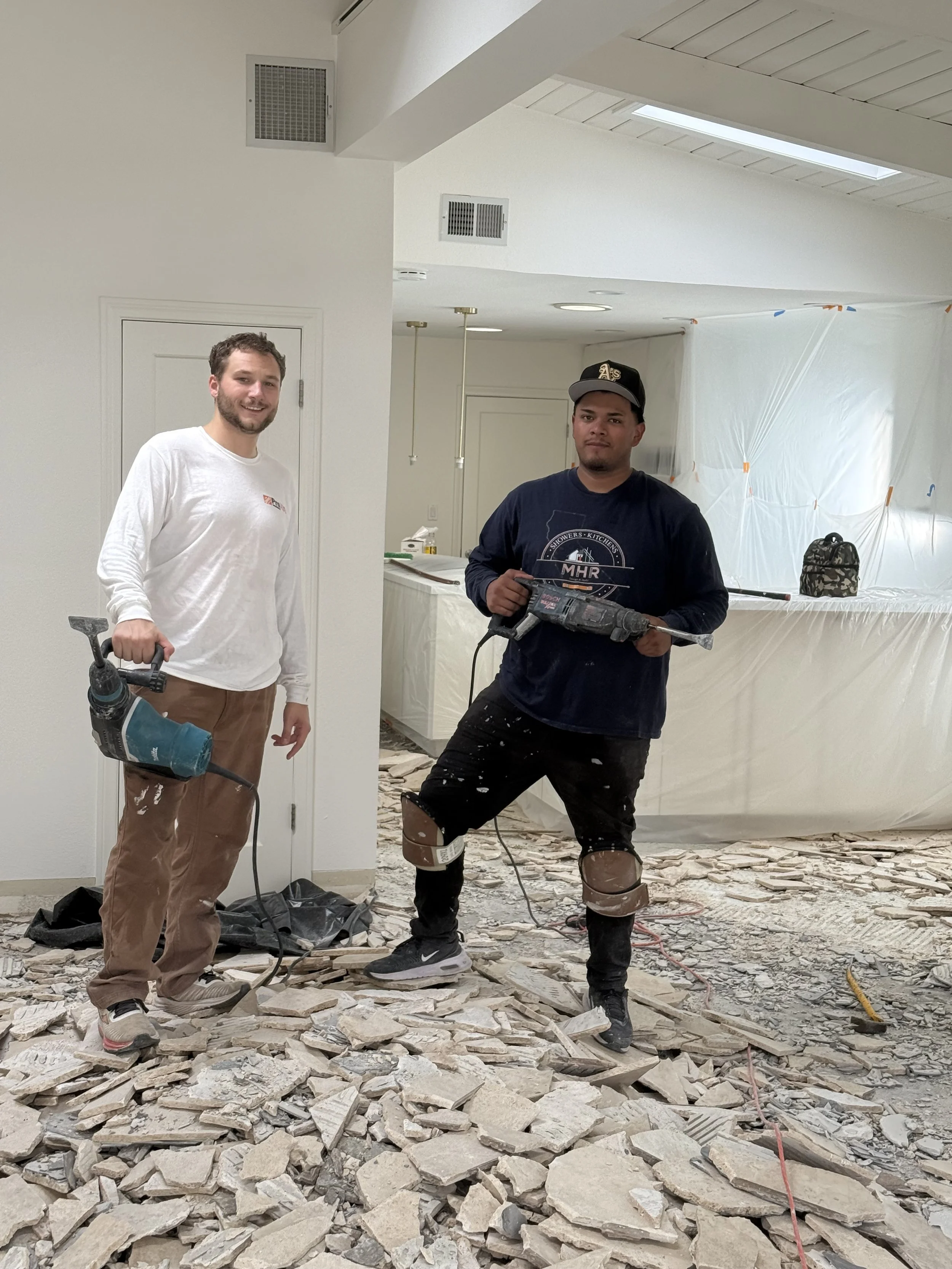Two construction workers standing on a debris-filled floor, holding power tools inside a partially renovated room, with plastic sheeting covering part of the background.