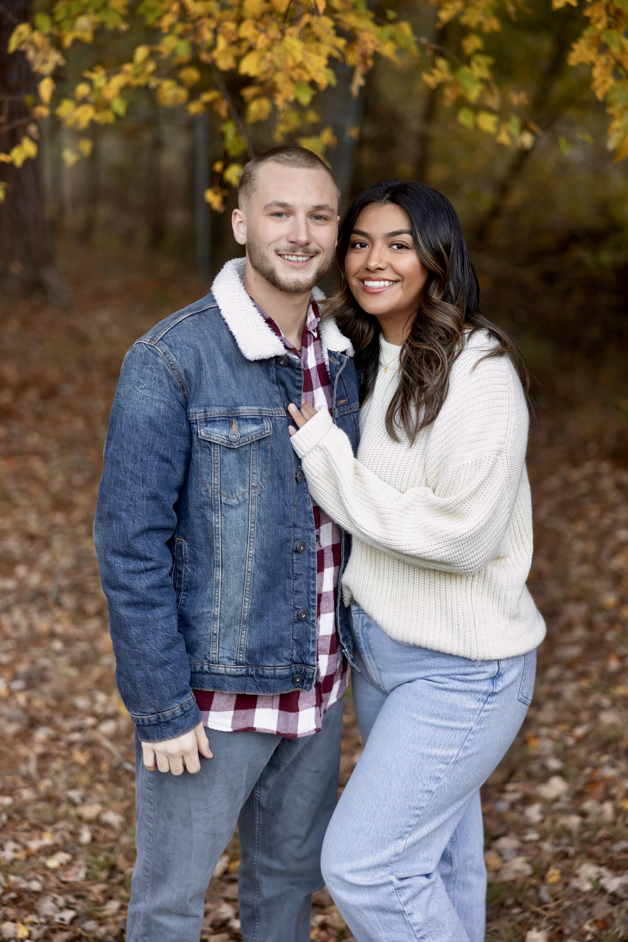 A smiling couple standing close together outdoors during fall, with colorful autumn leaves in the background.