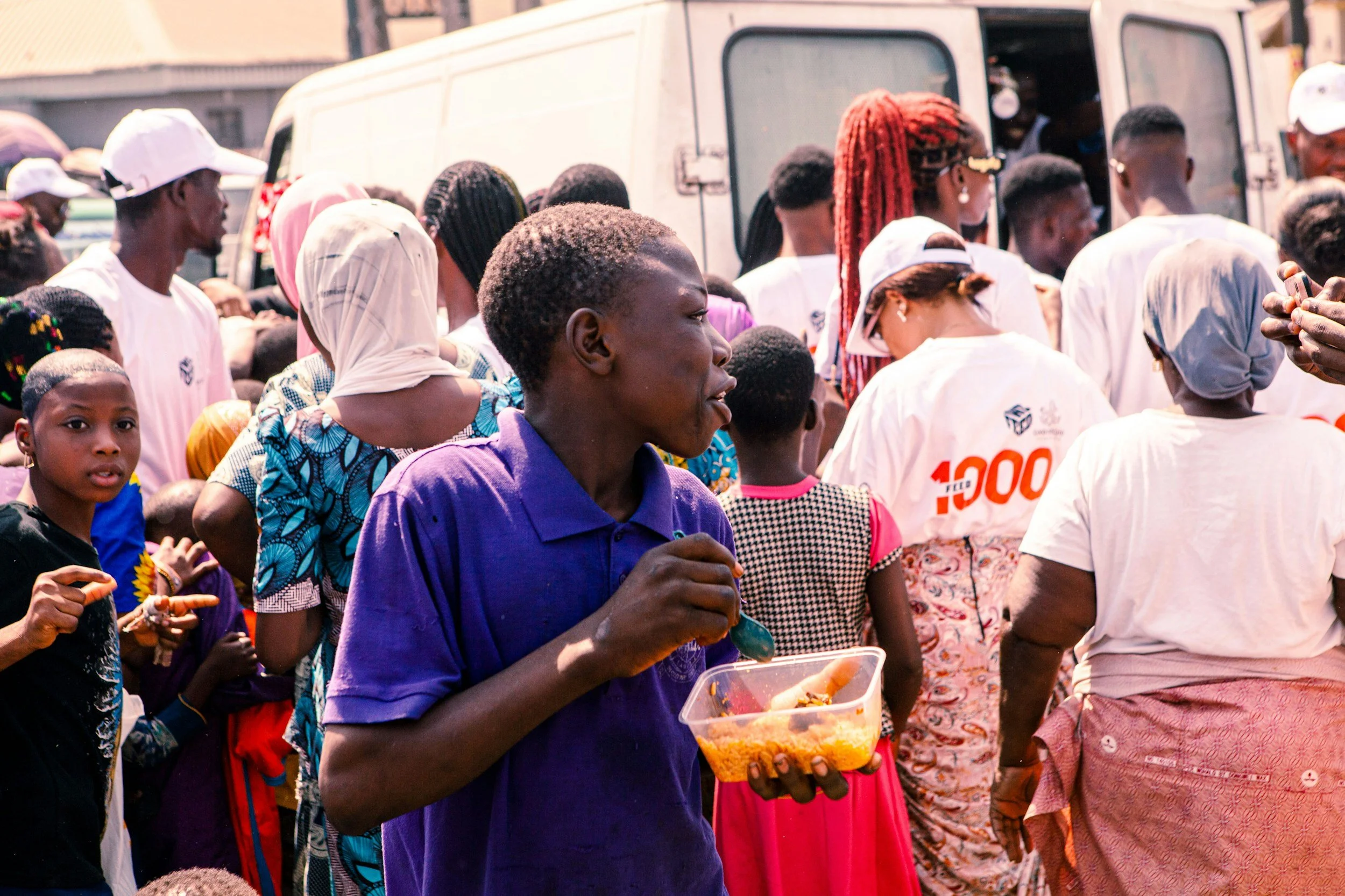 A crowd of people gathered outdoors, some wearing white shirts, some with headscarves, and a woman in a purple shirt holding a plastic container with food, standing near a white vehicle.