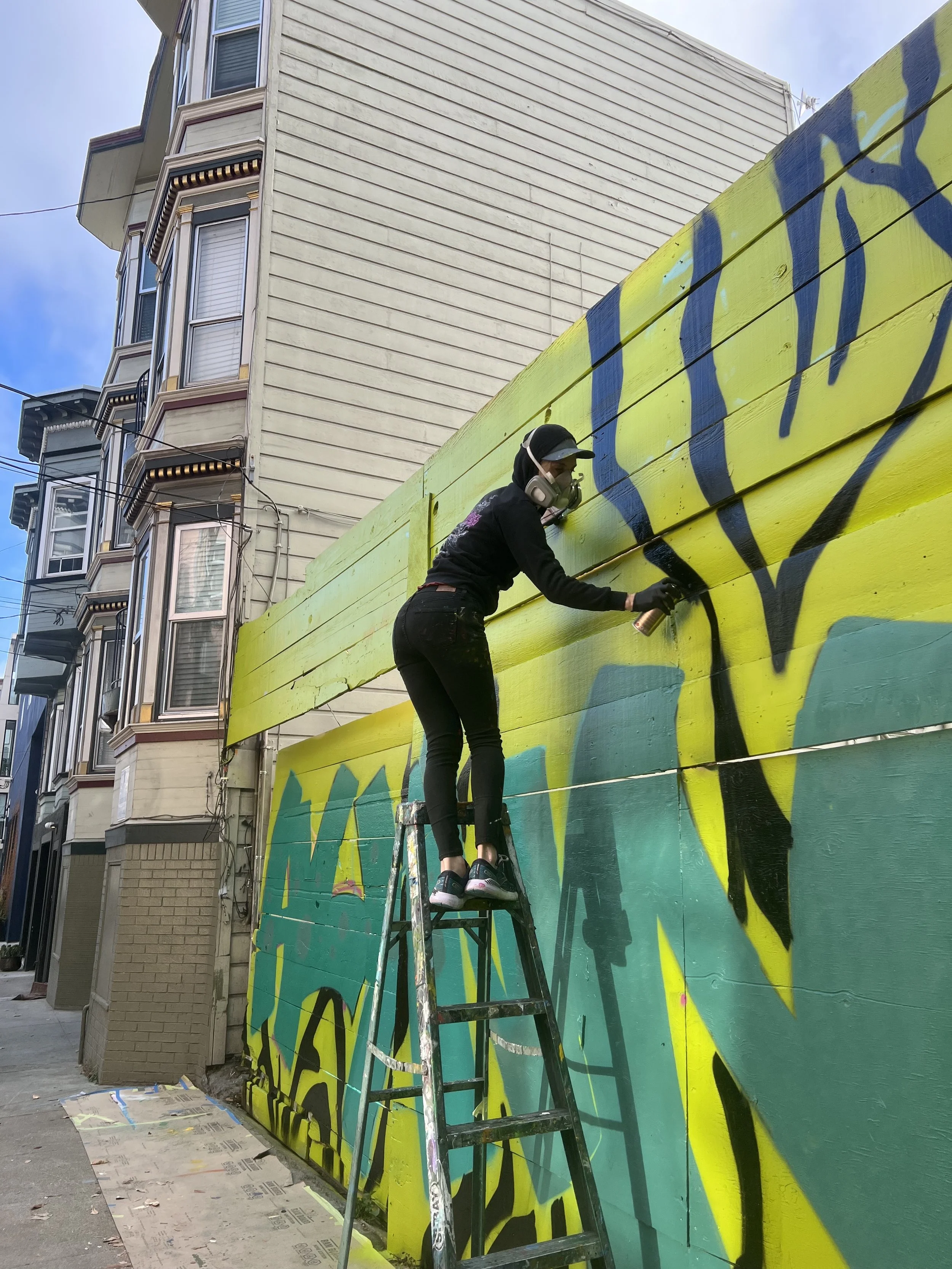 A person wearing a hoodie, mask, and gloves spray-paints graffiti on a large colorful mural on a wall outside a building, standing on a ladder.