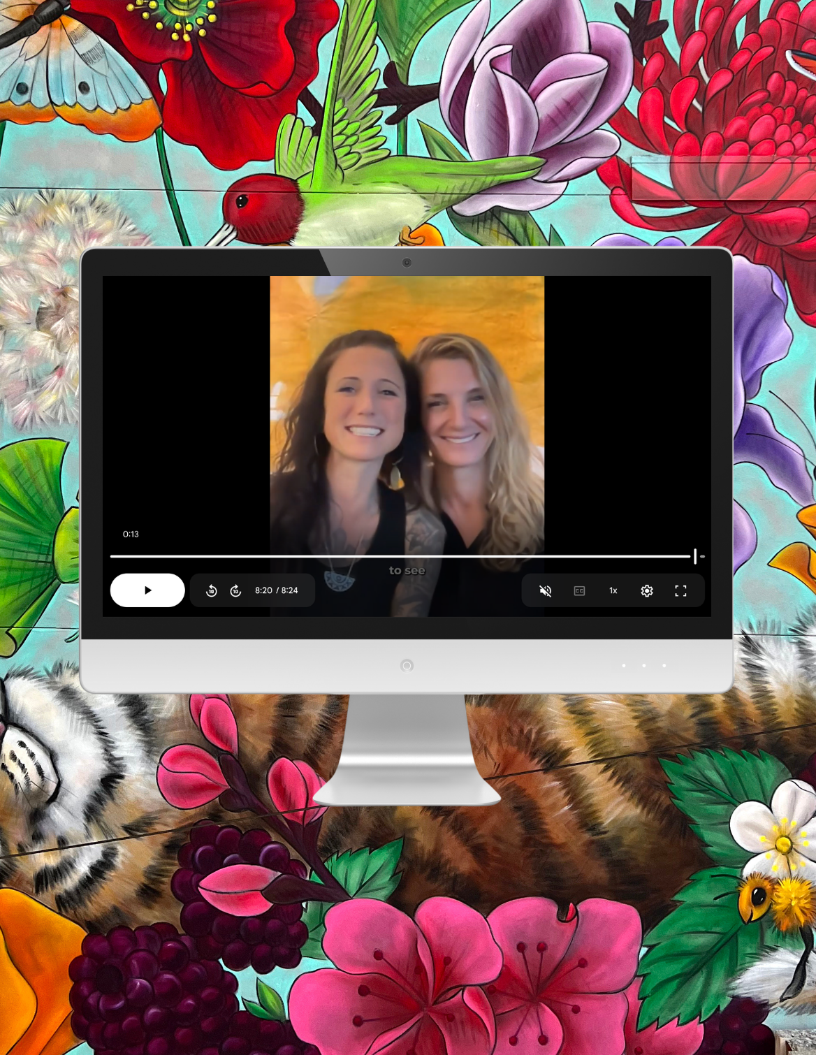 Computer monitor displaying a video with two smiling women, set against a colorful mural background with flowers and wildlife illustrations.