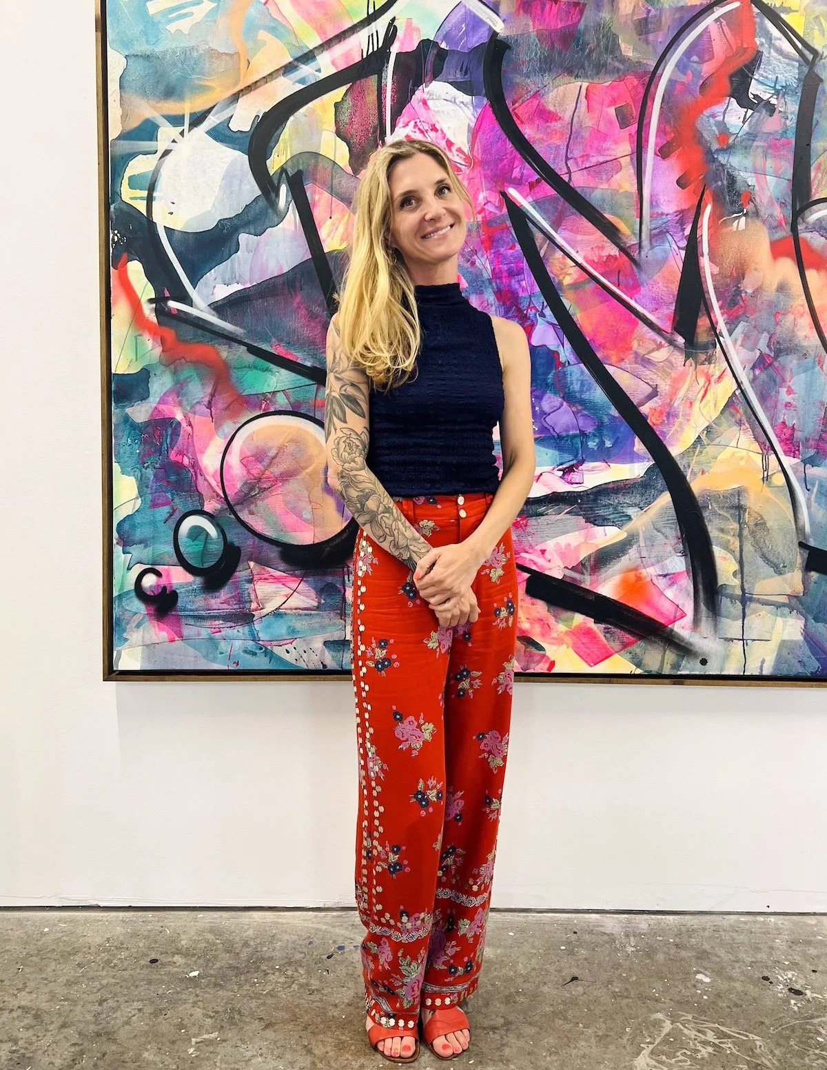A woman with long blonde hair, wearing a sleeveless navy top, red floral pants, and matching red sandals, stands in front of a colorful abstract painting at an art gallery.
