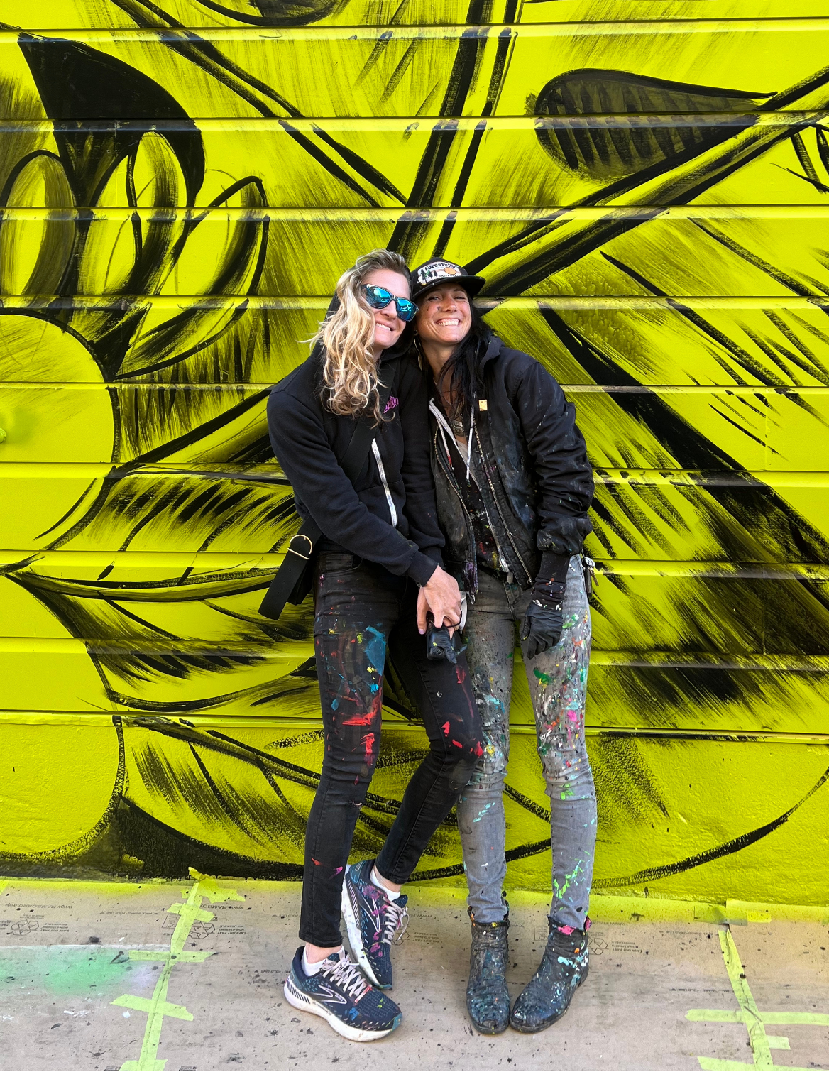 Two women standing in front of a large yellow and black street mural, wearing paint-splattered clothes and smiling.