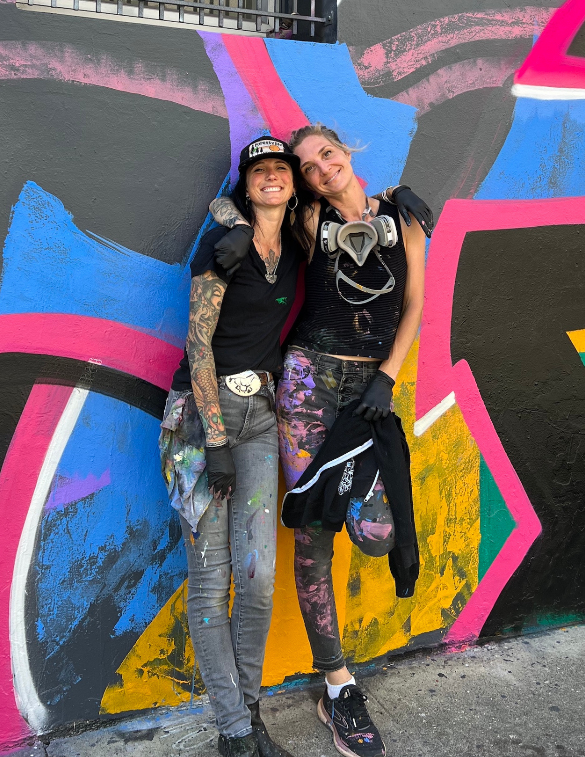 Two women smiling and standing close together in front of a colorful graffiti wall. One has tattoos on her arm, wearing a black t-shirt, gray pants, and a hat. The other has blonde hair, wearing a black top, patterned pants, and holding a black hoodie. Both are wearing gloves, with a respirator hanging around her neck.