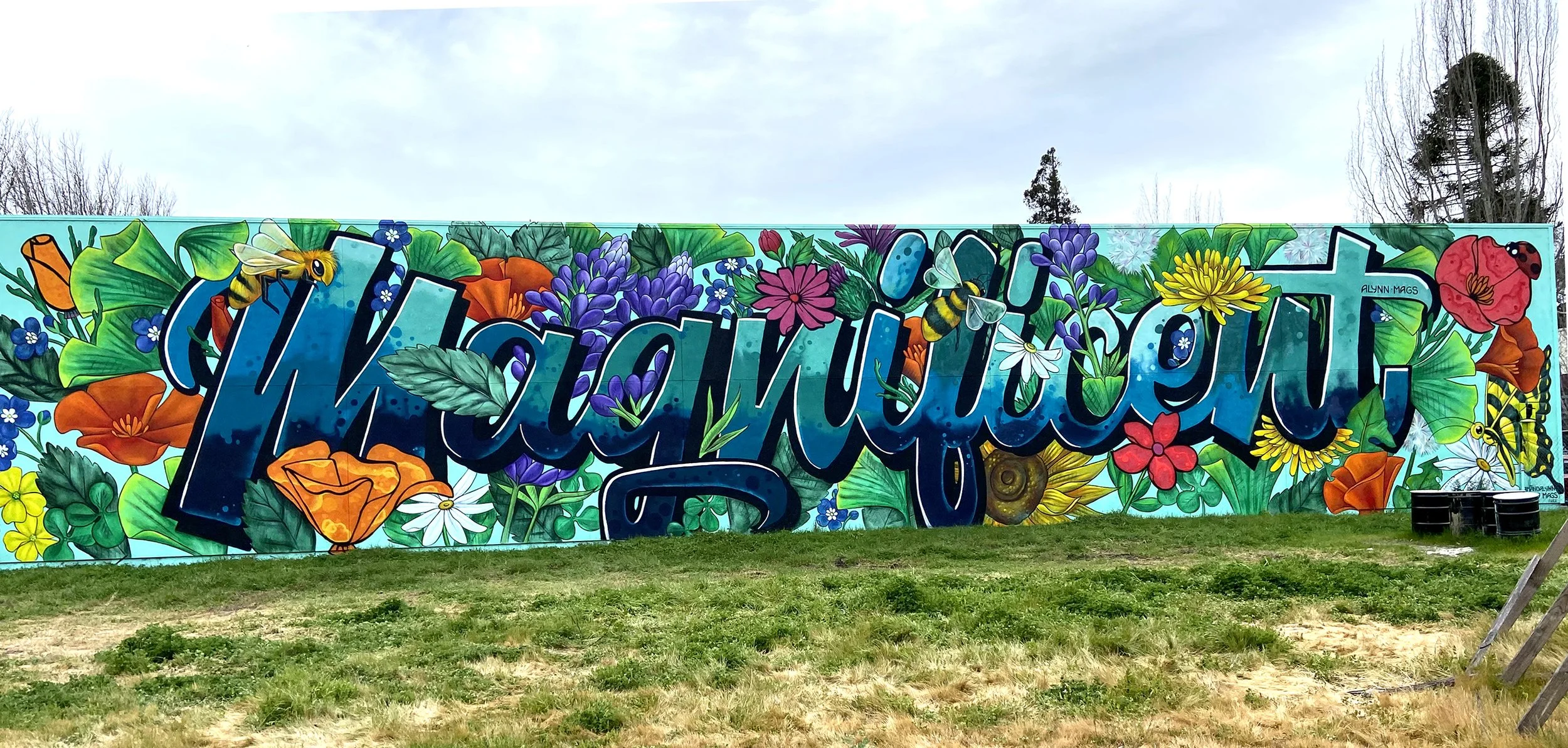 Colorful mural with the word 'Magnificent' surrounded by vibrant flowers, leaves, bees, and butterflies, painted on an outdoor wall.