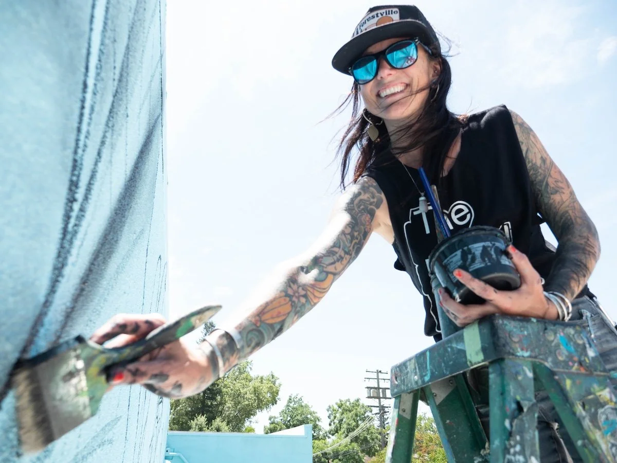 A woman with tattoos, wearing sunglasses and a cap, is painting a wall outdoors with a paintbrush, while smiling.