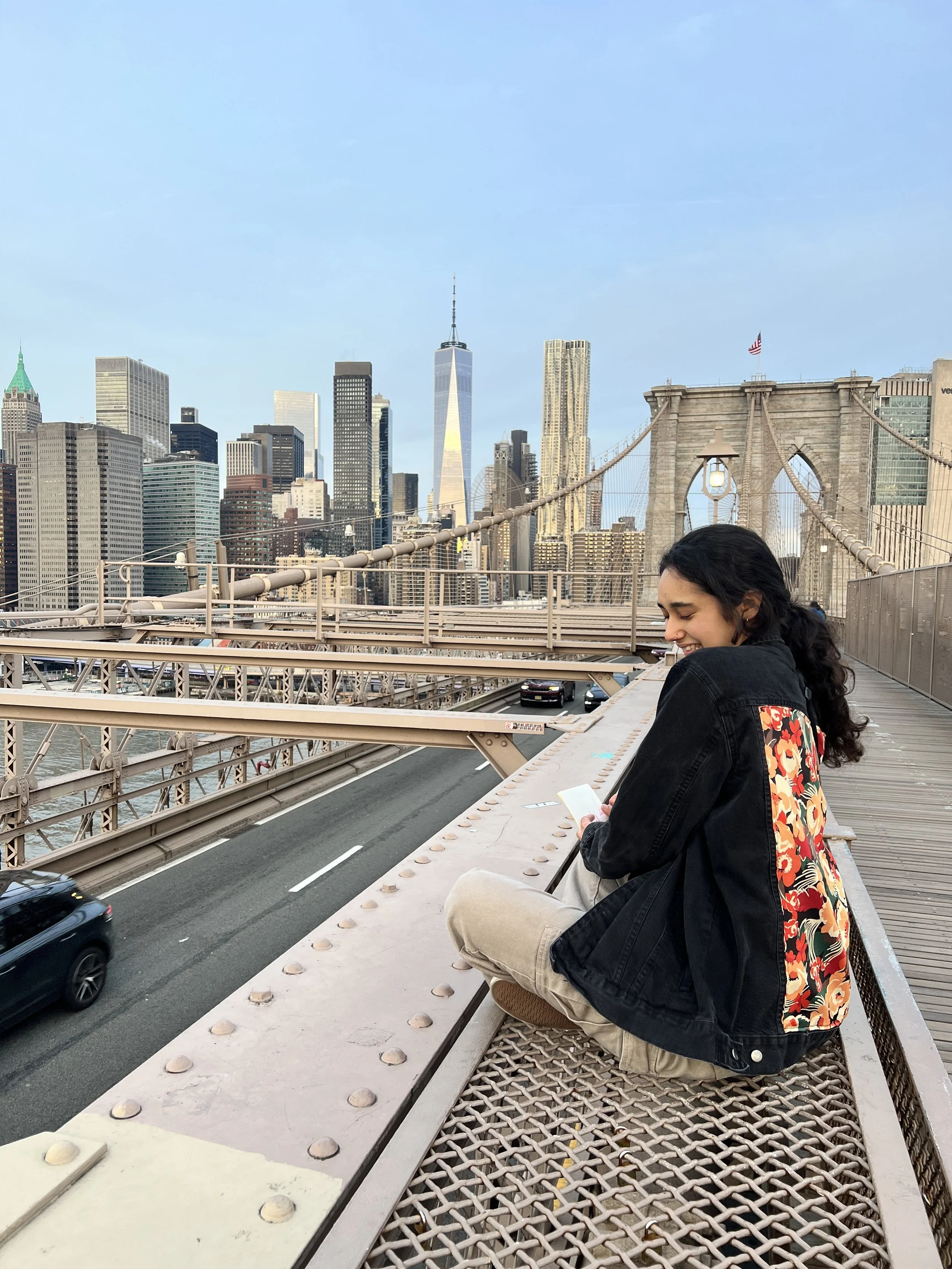 A woman with dark hair sitting on the Brooklyn Bridge in New York City, smiling and looking at her phone with the Manhattan skyline, including One World Trade Center, in the background.