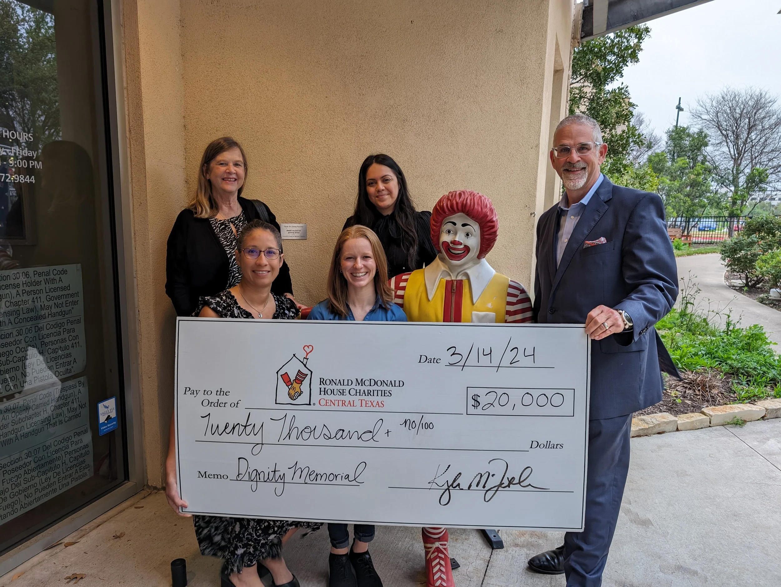 A group of six people and Ronald McDonald holding a large check for $20,000 made out to Dignity Memorial, dated 3/14/24, standing outside a building with trees in the background.