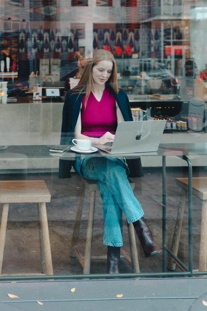 Woman with long red hair working on a laptop inside a coffee shop, sitting at a table with a cup of coffee and a smartphone, seen through a glass window.