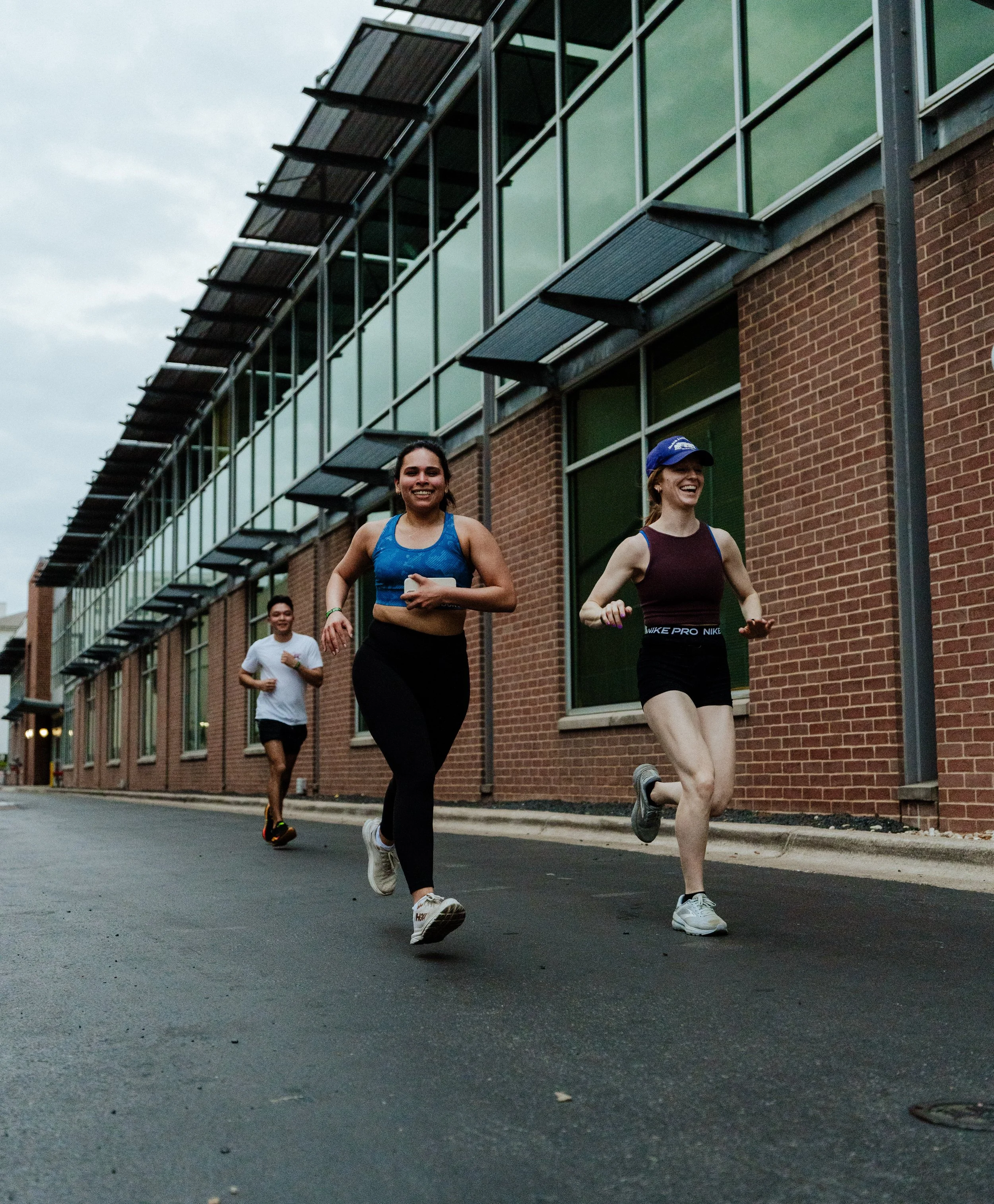 Three people jogging on a city street near a brick building with large windows.