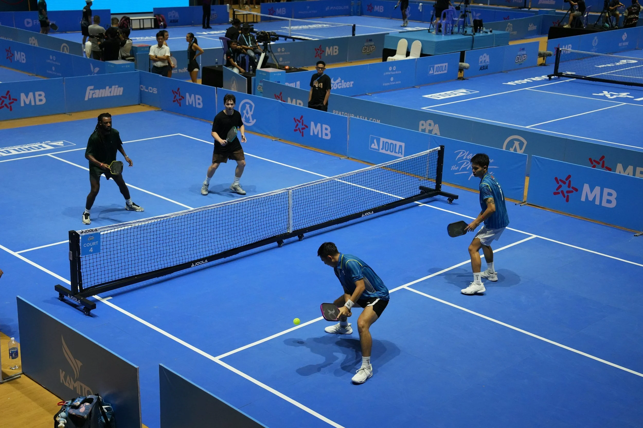 Two players are practicing pickleball with paddles on a blue court, while two opponents are positioned near the net, ready for the game, with a referee and other players visible in the background. Enjoy tournament play at Auckland Pickleball Centre.