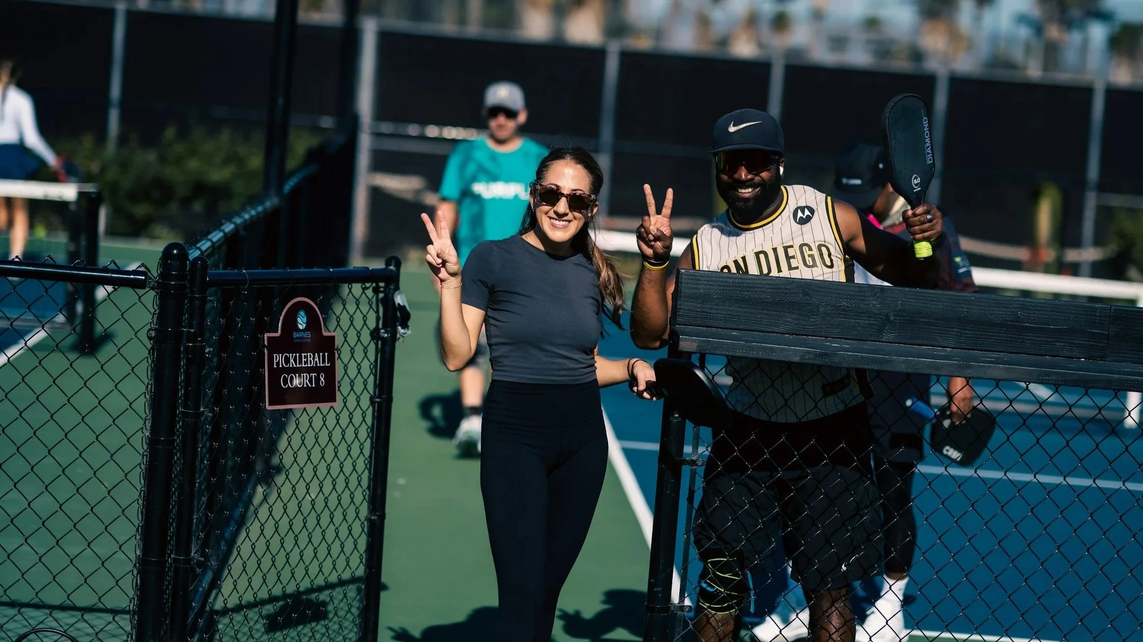 Two people on a pickleball court making peace signs, smiling, and holding paddles, with others in the background. See Membership options at Auckland Pickleball Centre.