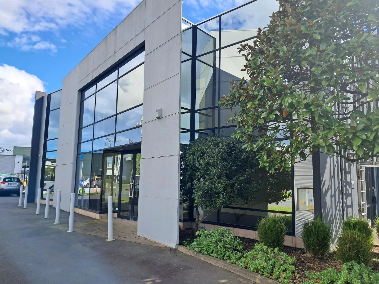 Auckland Pickleball Centre's modern main commercial building with large glass windows, surrounded by landscaping including trees and bushes, and a parking area with cars, under a partly cloudy sky.