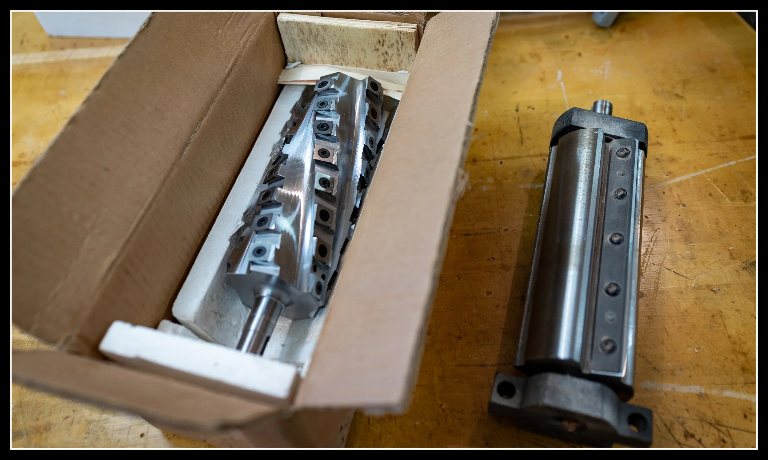 Two jointer cutterheads on a workbench: original Steel City straight‑knife head beside a new carbide helical head, shown during a woodworking machine upgrade.