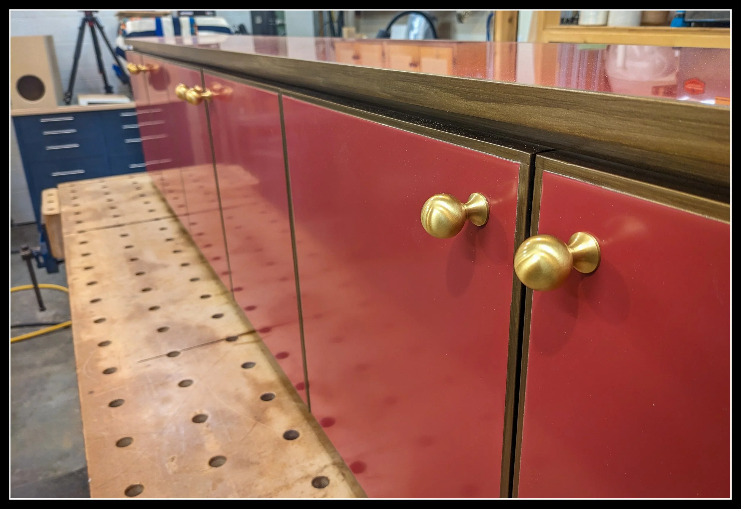 Gloss‑red cabinetry with walnut trim and brass knobs, photographed in a woodworking shop with a workbench in the foreground.