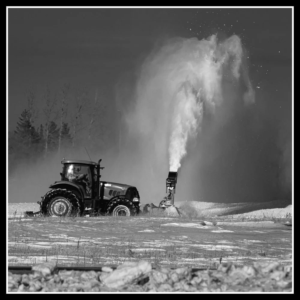 A tractor blowing snow high into the air on a prairie winter day, capturing the cold outside the future hearth room.