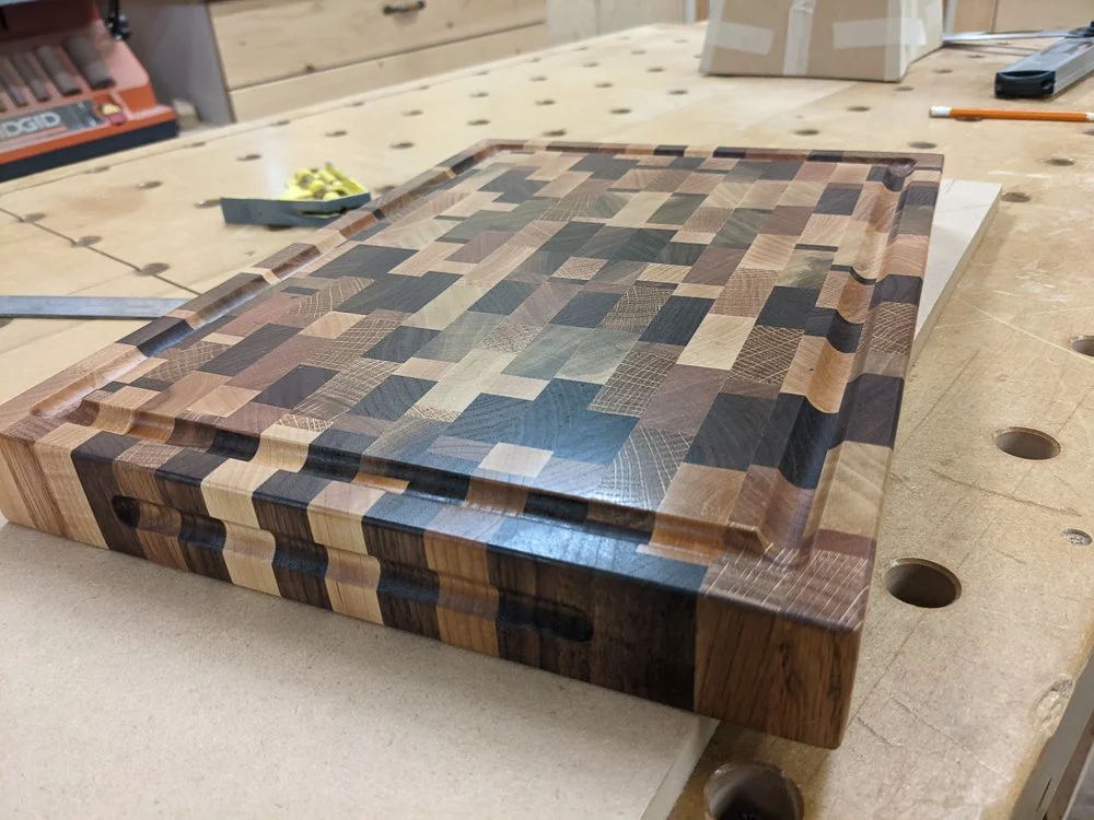 End‑grain cutting board showing mixed hardwood species from various projects, with juice groove and polished edges.