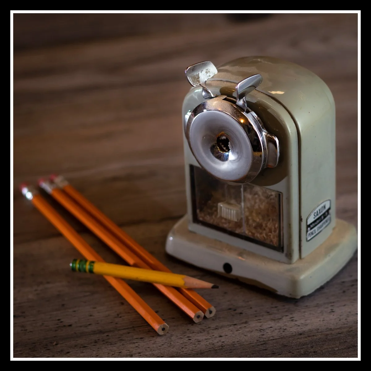 Vintage Saxon desk pencil sharpener with yellow HB pencils on a wooden surface, showing metal faceplate and shavings box.