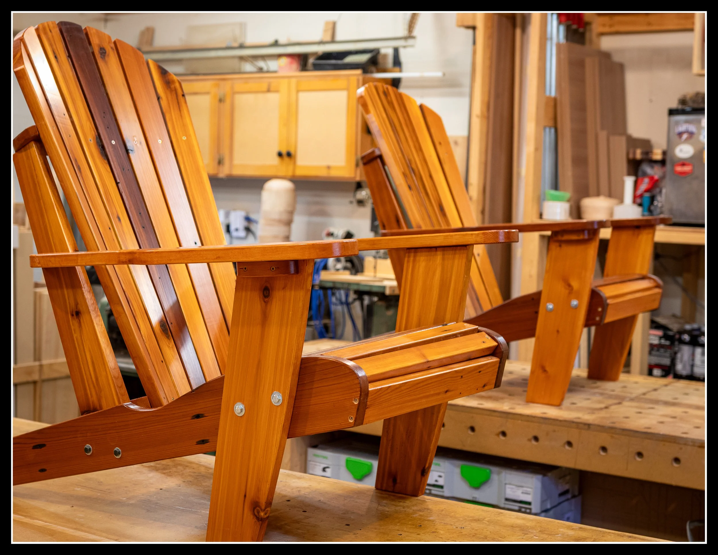 Two finished cedar Muskoka chairs in a woodworking shop, built from reclaimed deck boards and polished to a warm, rich glow.