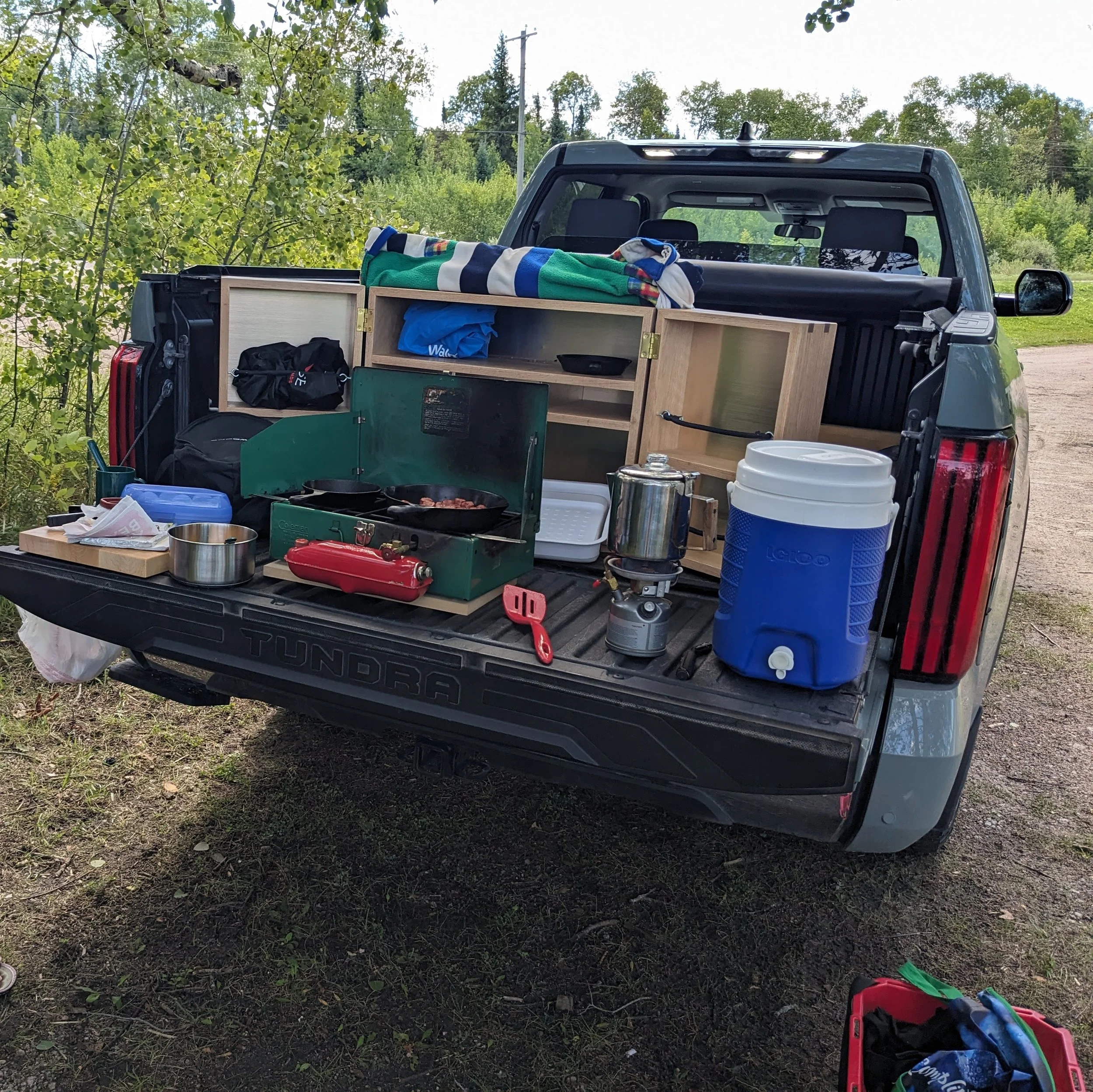 Tailgate cooking setup with restored Coleman stove, percolator, water cooler, and Mini Kitchen cabinet during a truck‑nic.