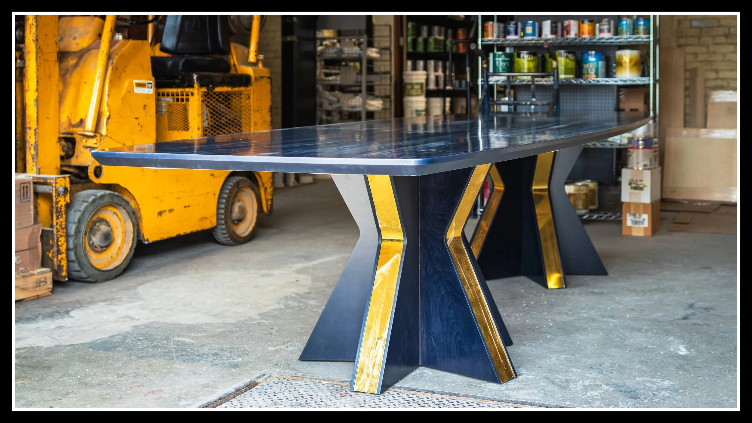 12‑foot solid maple dining table with dark stain, 2K poly finish, and dual five‑point star pedestals featuring brass‑colored inlay, shown in a workshop setting.