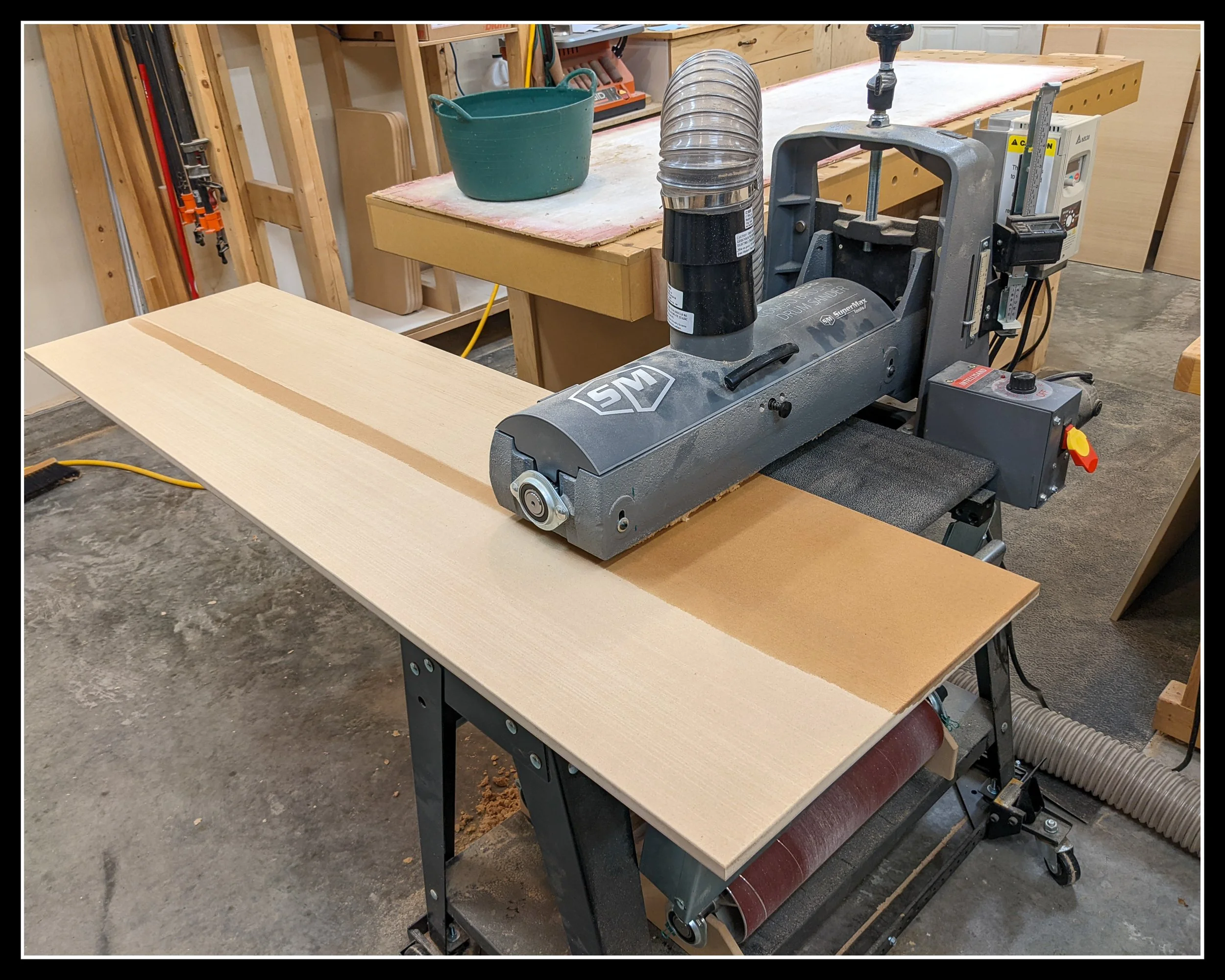 SuperMax drum sander in a woodworking shop with a board running through it, showing a compact machine that requires careful feed monitoring for smooth, consistent sanding.
