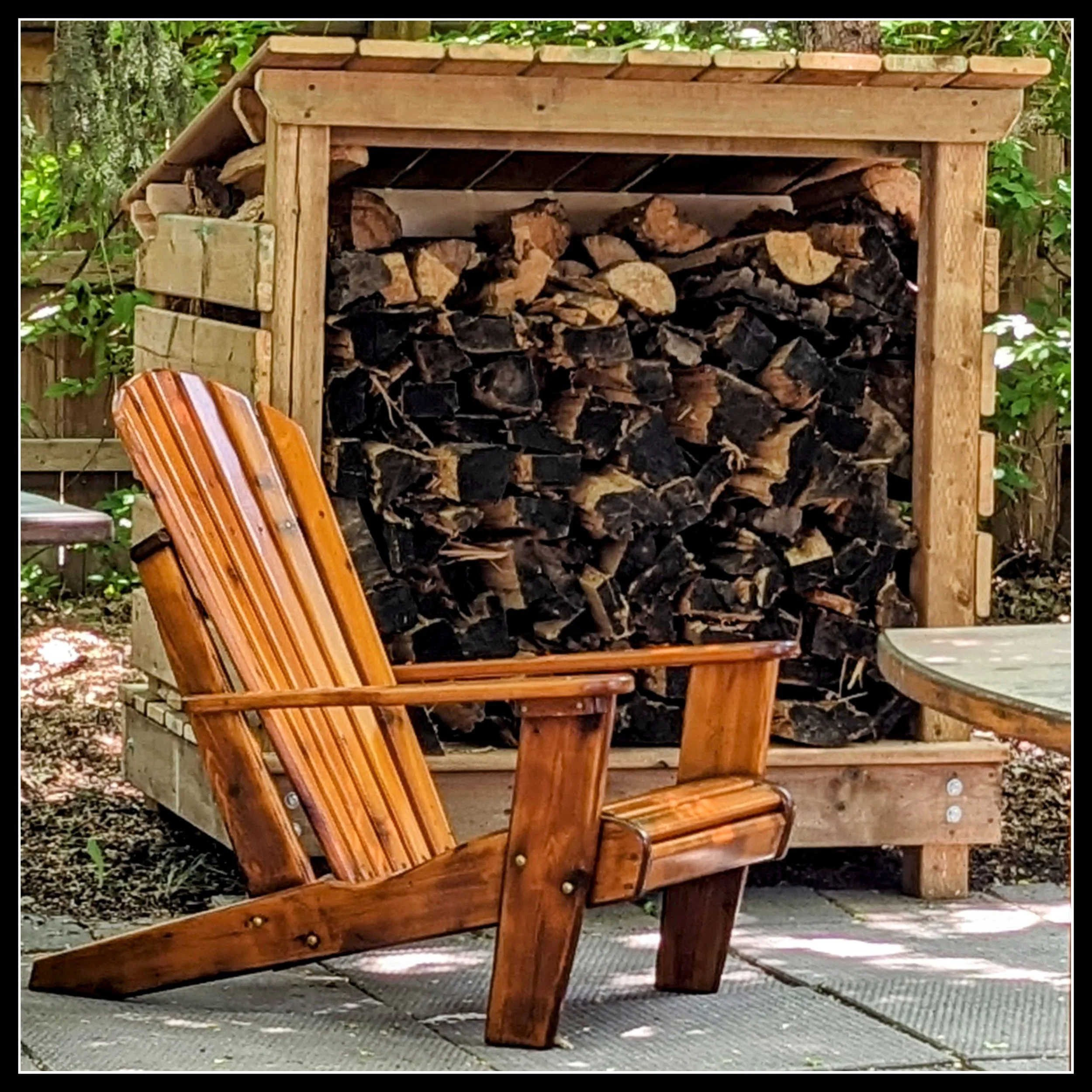 A small three‑walled backyard firewood shelter with a lean‑to roof, filled with weathered poplar firewood showing dark end grain; trees surround the area and a cedar deck chair sits in the foreground, with the shed as the main focus.