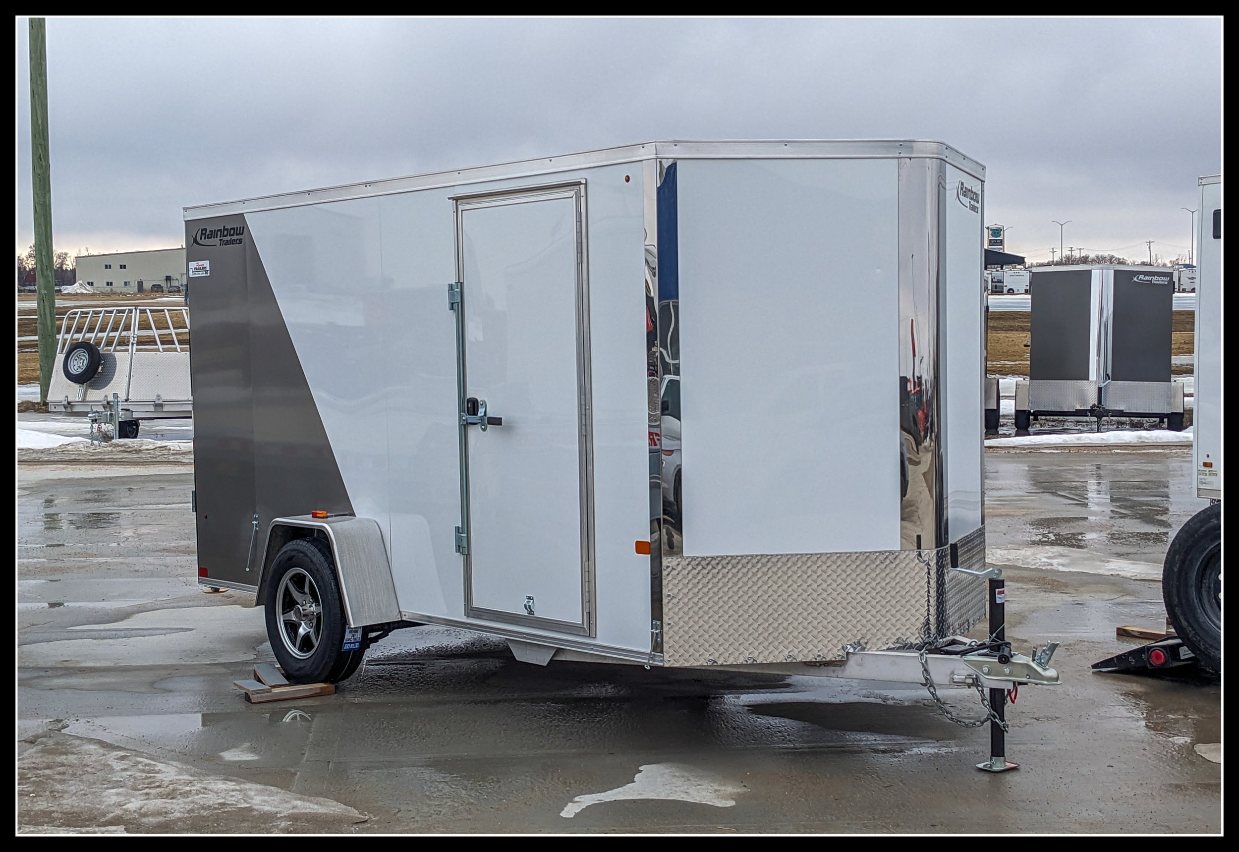 2024 Rainbow enclosed 12’ cargo trailer with single axle, white and gray panels, side door, and diamond‑plate front, parked on wet pavement in a storage lot.