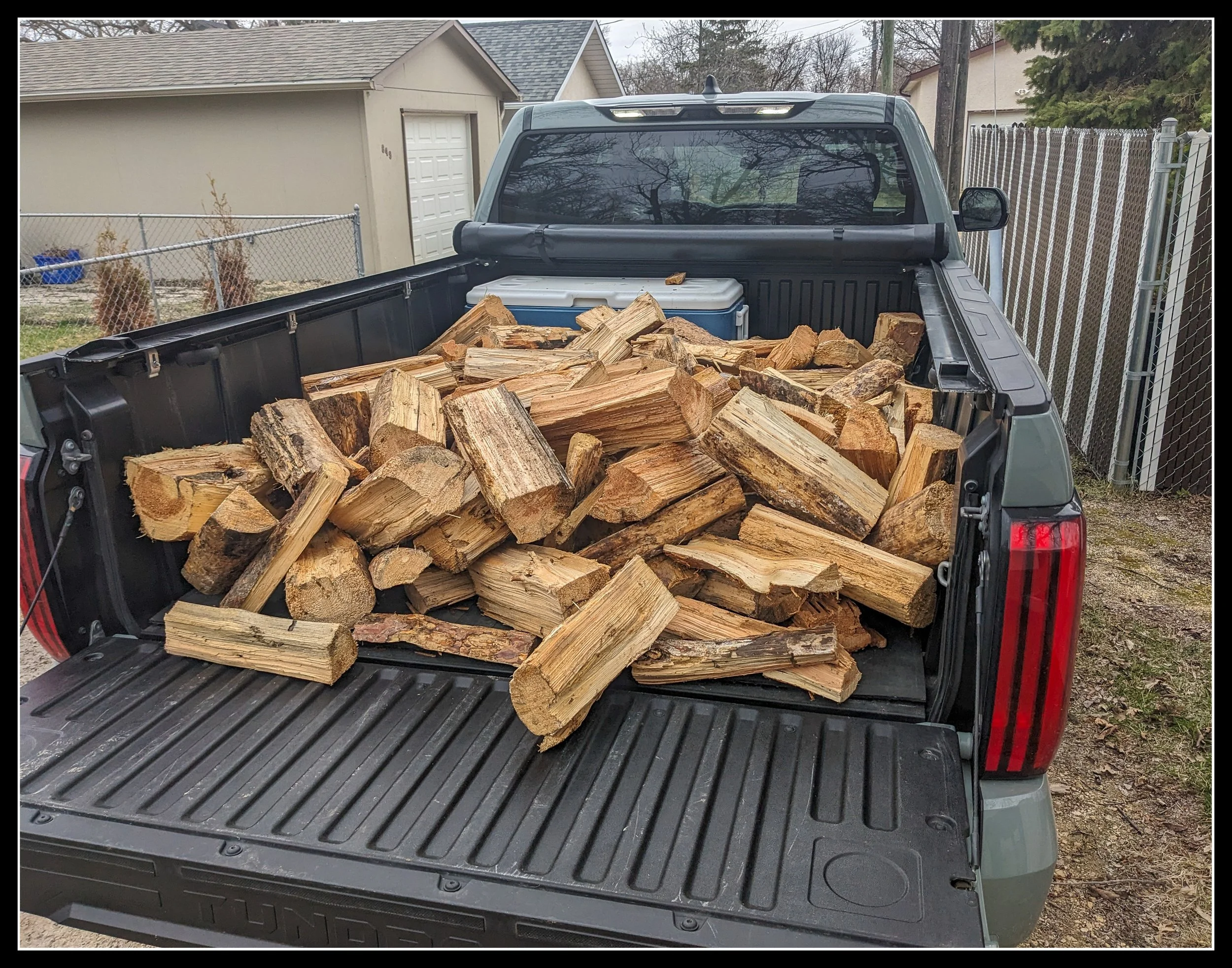 Back view of a pickup truck loaded with split pine firewood in a back lane on a cloudy day, showing rough logs stacked in the bed.