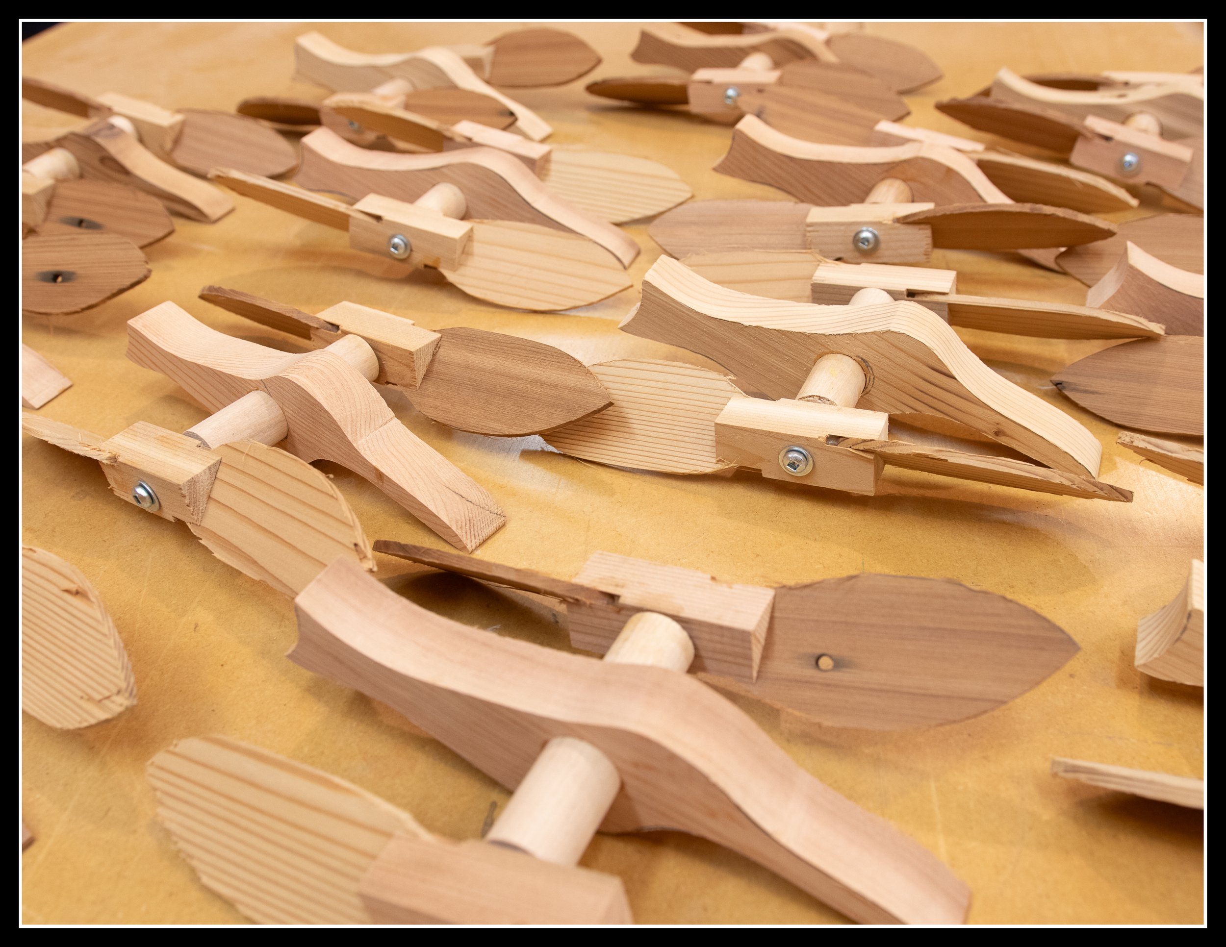 A set of handcrafted cedar whirlybird parts arranged on a workbench, showing curved wooden arms, dowels, and pivot hardware — a simple, kid‑friendly woodworking project made with full supervision.