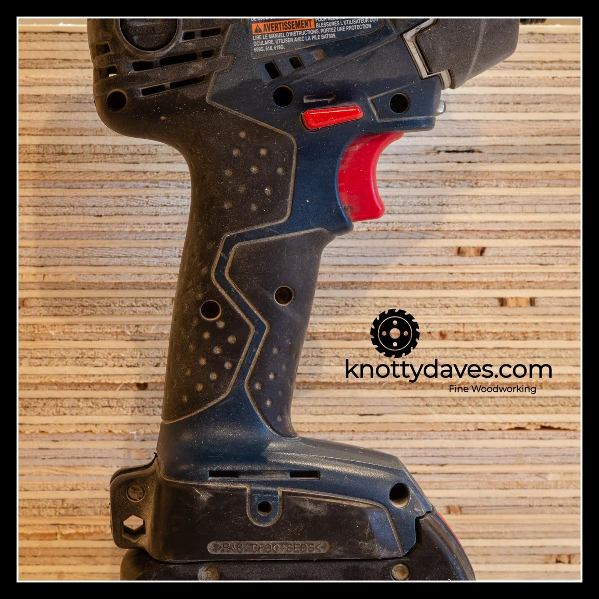 Heavily used Bosch 18V cordless drill with worn grip and red trigger, photographed against stacked plywood; fifteen‑year‑old tool showing durability after multiple battery replacements and years of job‑site abuse.