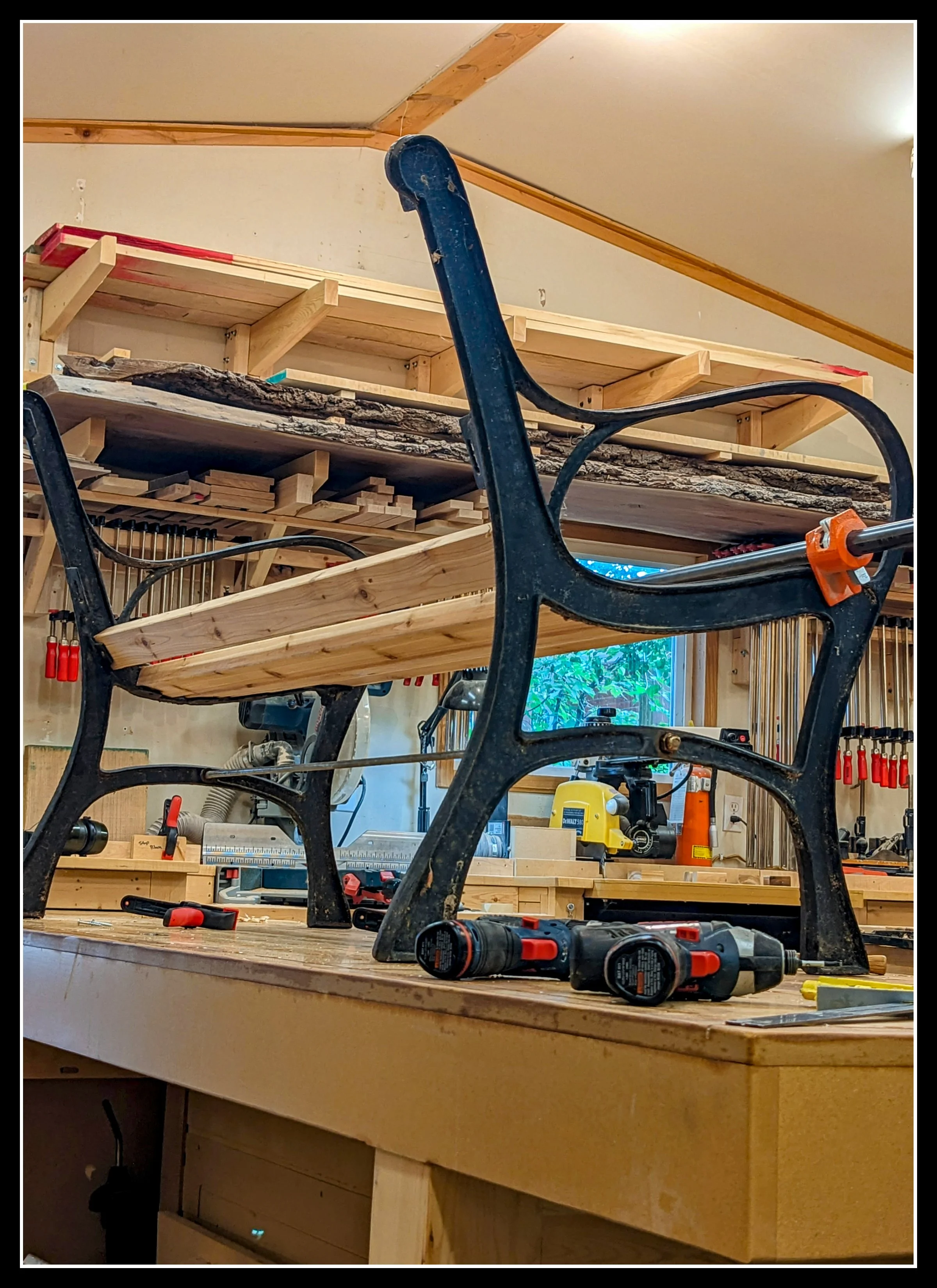 View of a woodworking workshop with a iron and cedar garden bench on a wooden workbench. The chair's seat is replaced with a new wooden frame,  Various tools, including cordless drills, are scattered on the workbench. In the background