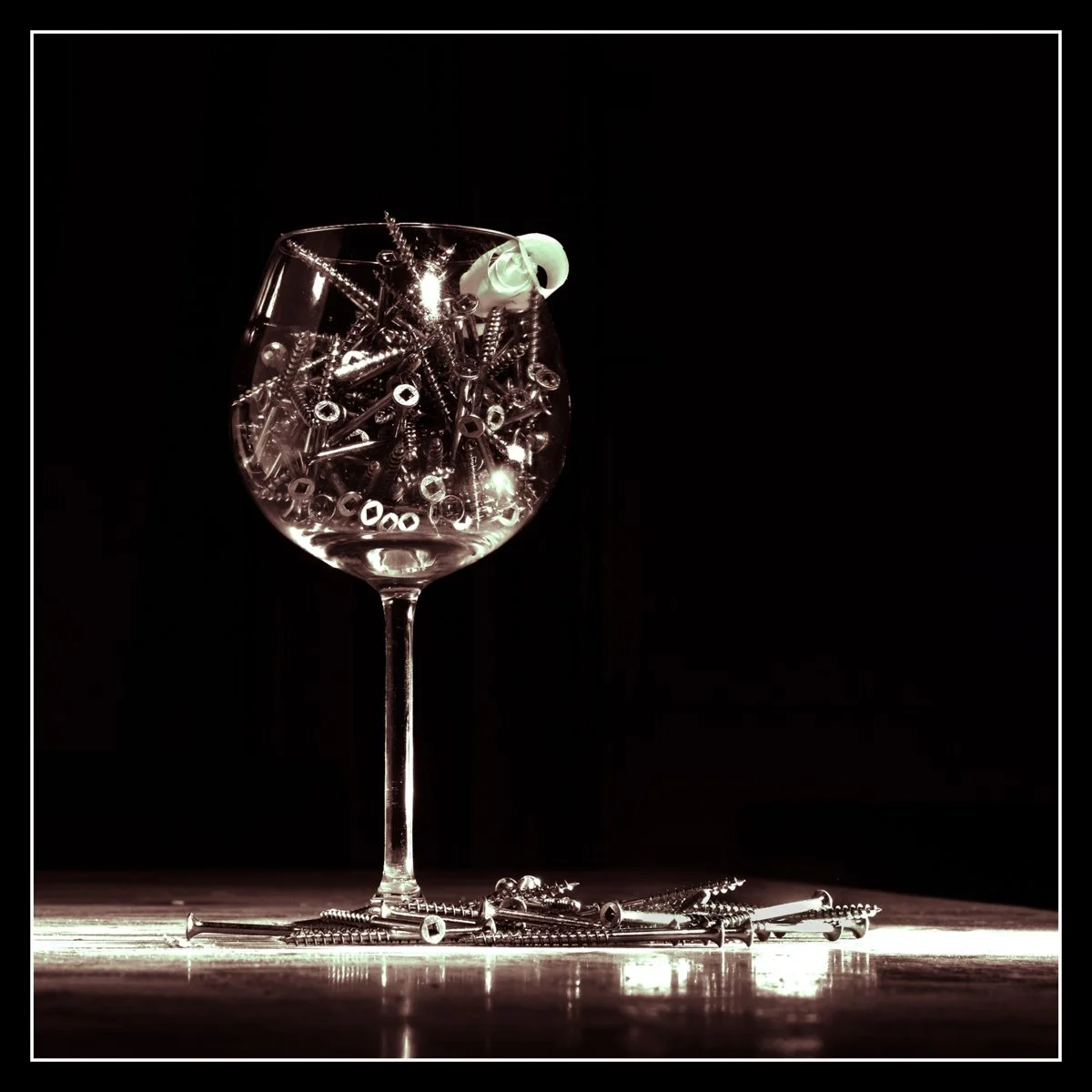 Wine glass filled with wood screws on a glossy surface, lit with overhead and side flashes, with a single matte hand‑plane ribbon curl resting on the rim.