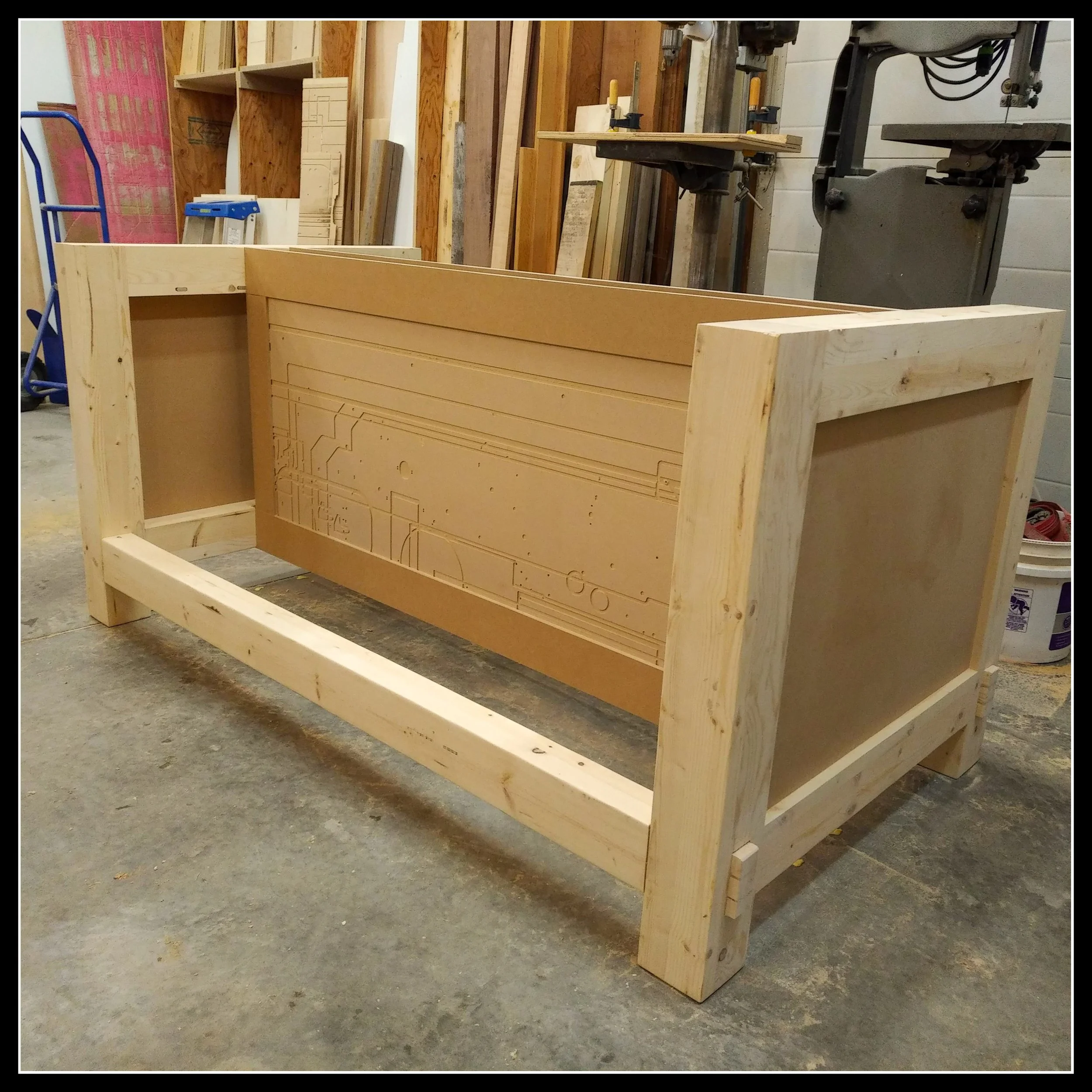 A heavy, square MFT workbench under construction in a modern woodworking shop, with a framed top and tools in the background.