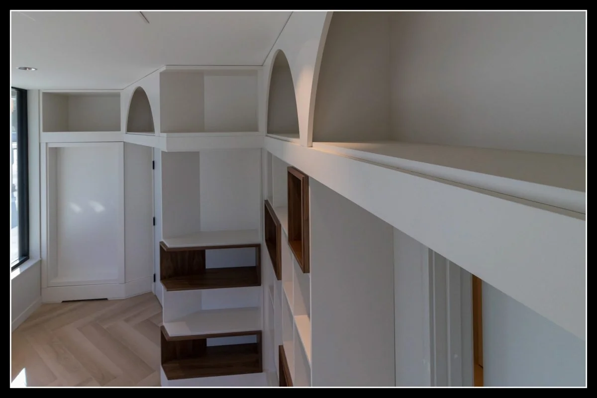 White built-in shelving with arch-shaped compartments and open cubbies in a room with a window and hardwood flooring.