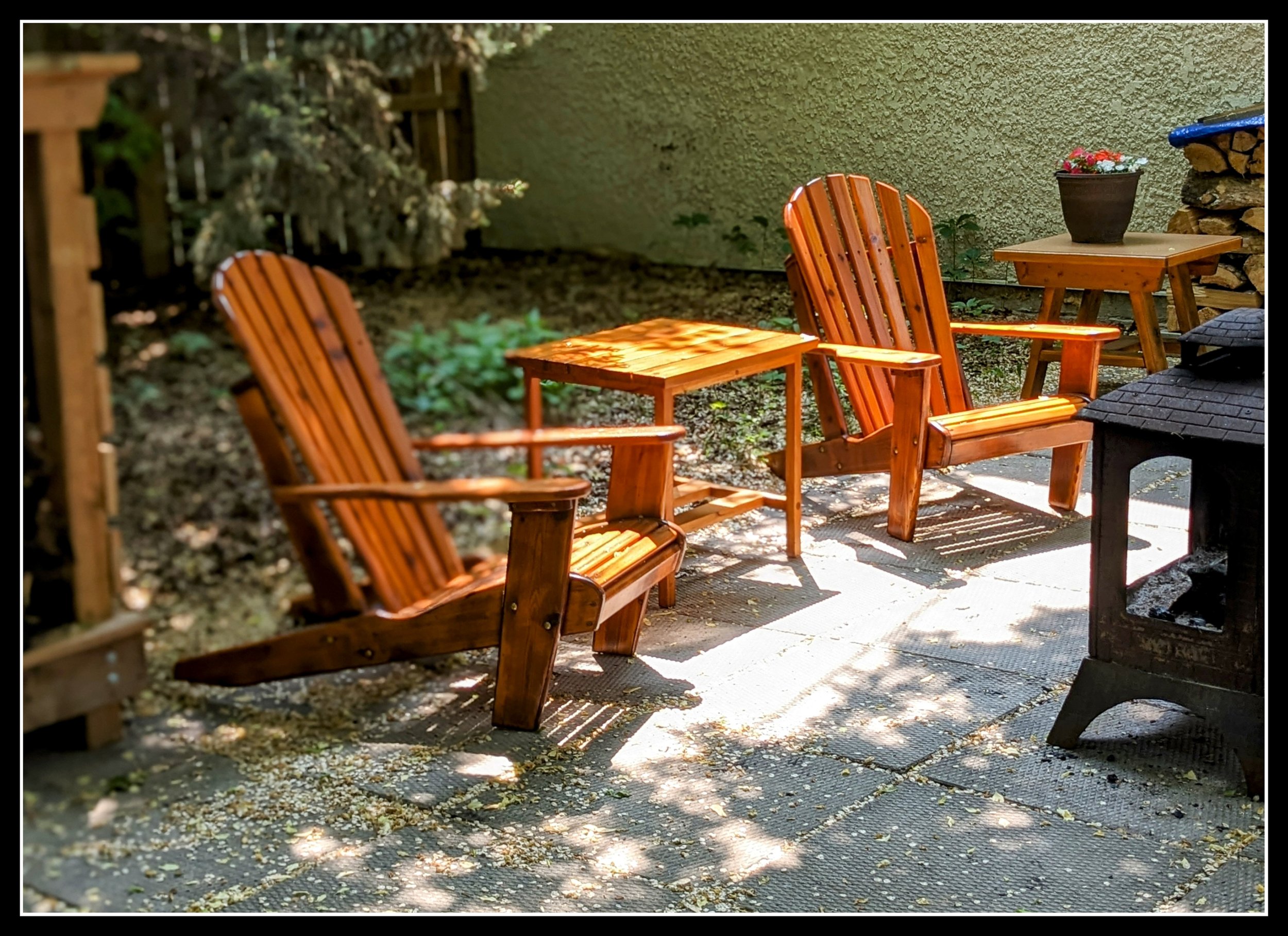 Two wooden Adirondack chairs with a matching small side table in an outdoor garden setting, with sunlight filtering through trees, a potted plant on a wooden table, and a storage stack of firewood in the background.
