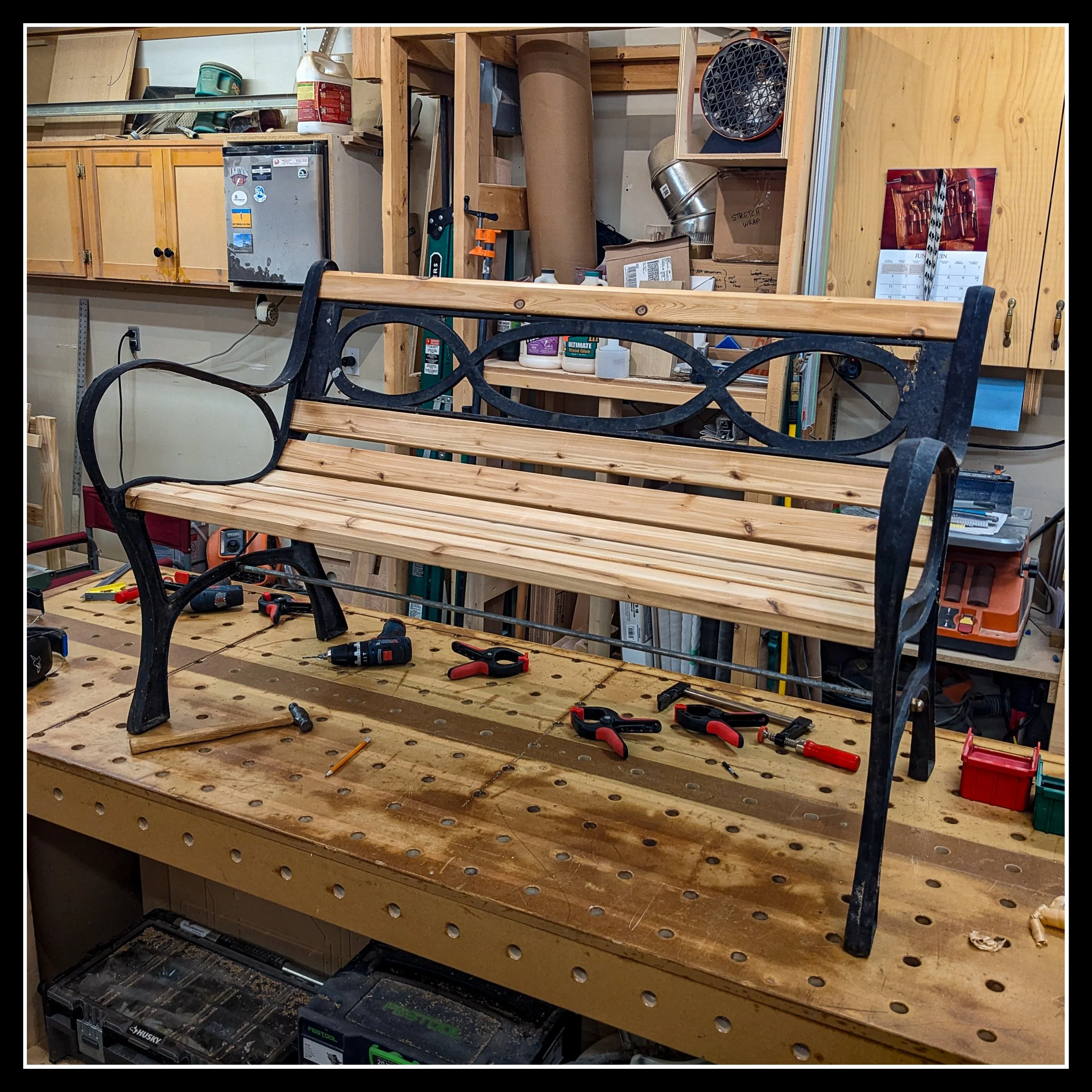 Wooden and metal garden bench being assembled in a woodworking shop, with tools and supplies on the workbench and shelves in the background.