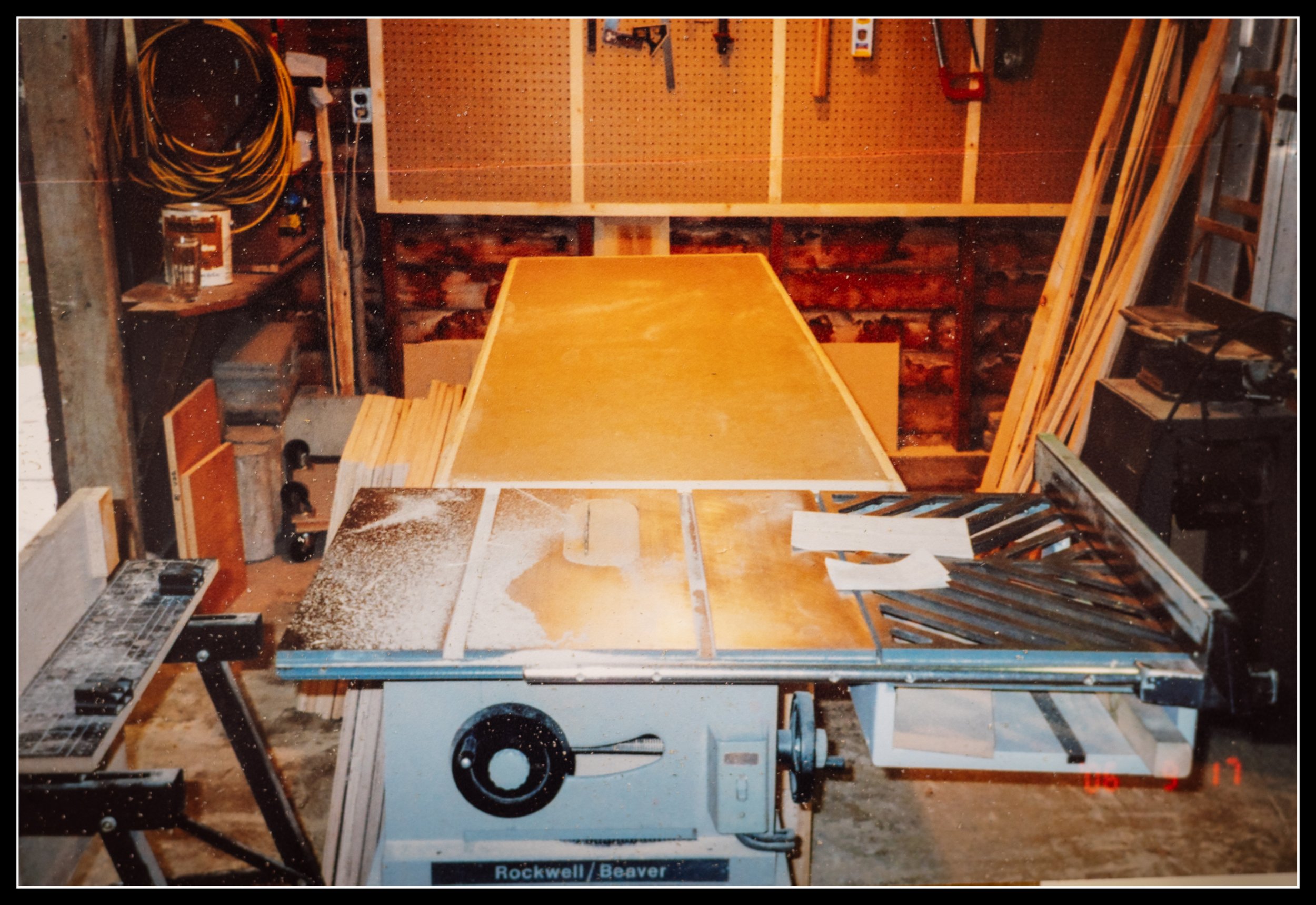 A Rockwell Beaver contractor table saw in a woodworking shop, covered in sawdust, with plywood on a bench behind it, tools hanging on the wall behind it and lumber stacked along the wall. A workshop showing the early tools that shaped the woodworker.