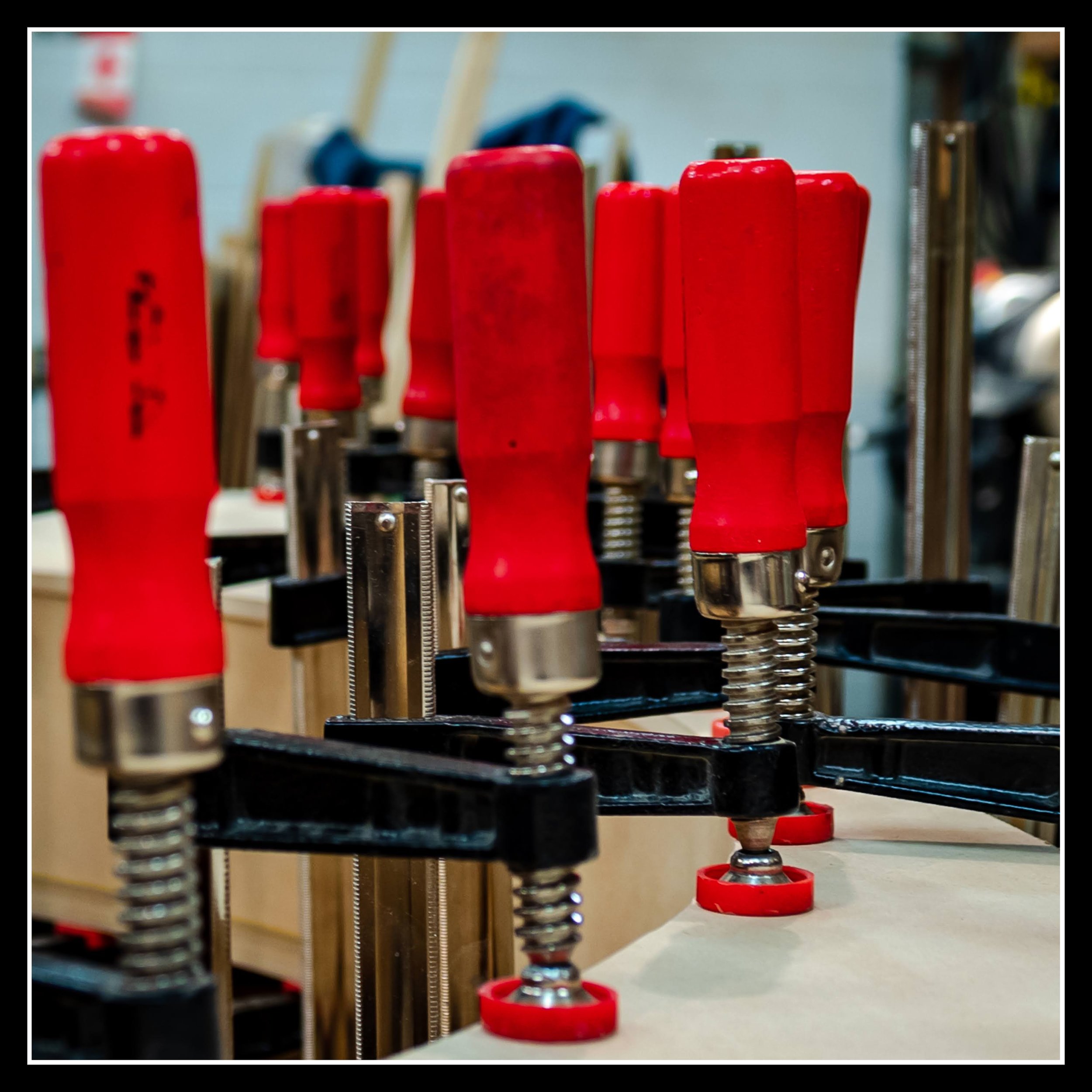 Benchmark F‑clamps securing boards together in a woodworking shop, red handles visible, holding wood steady during a glue‑up.