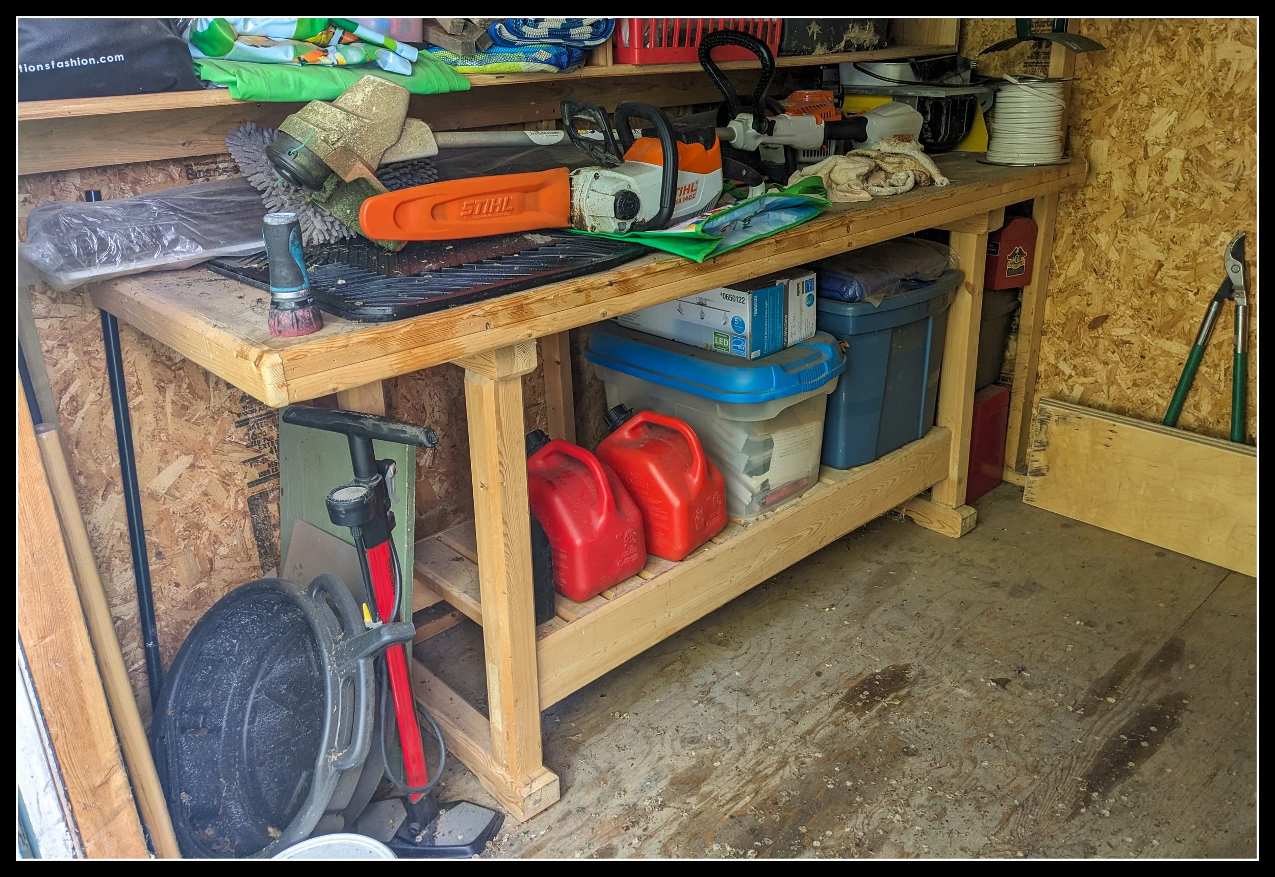 Old spruce workbench in a wooden shed, showing hand‑worn surfaces, tool marks, and a STIHL chainsaw with fuel cans and woodworking tools stored underneath.