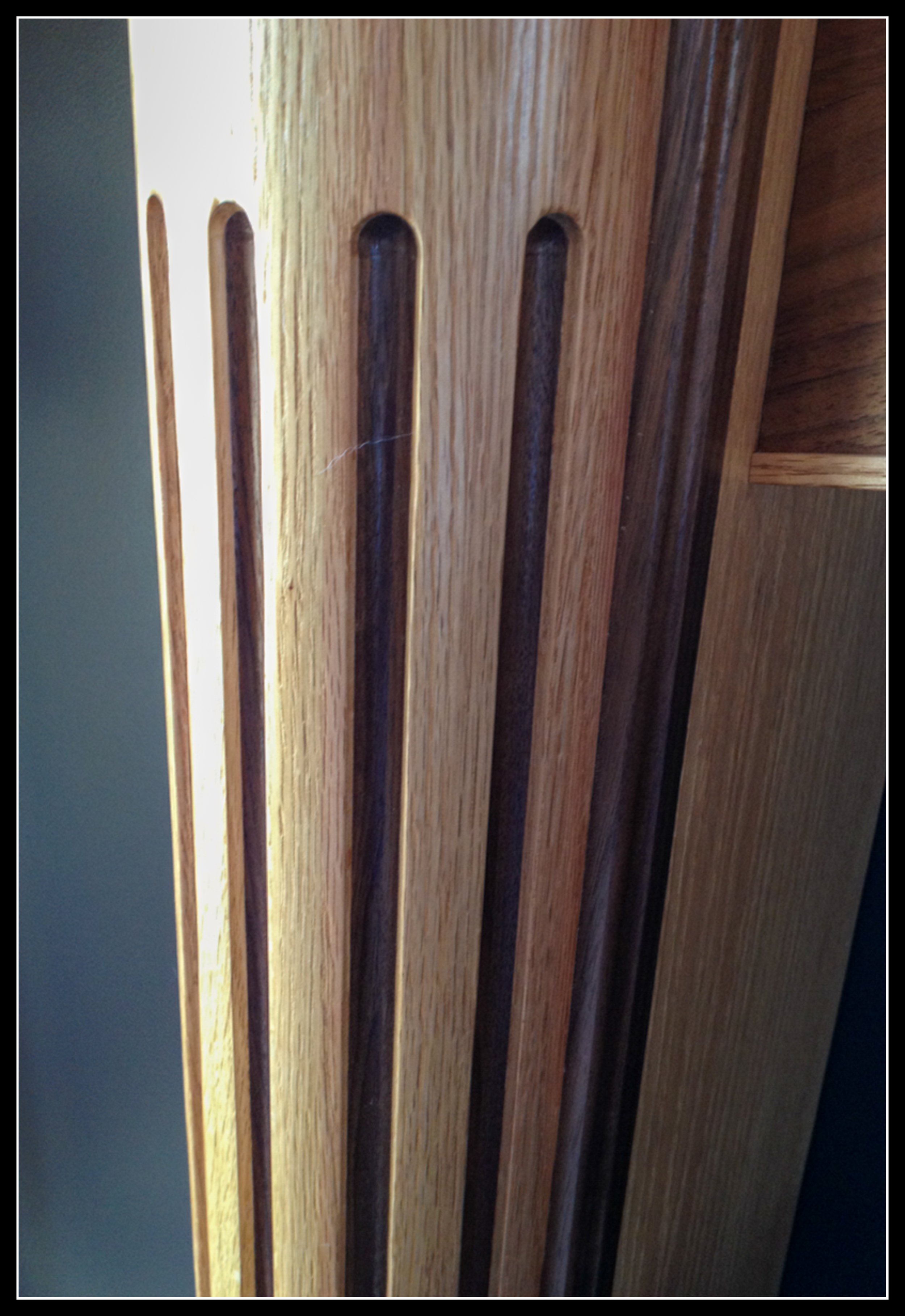 Close‑up of a mantle column showing a solid walnut inlay set into rift‑cut red oak with a clear natural finish, carved with vertical grooves and detailed woodworking craftsmanship.
