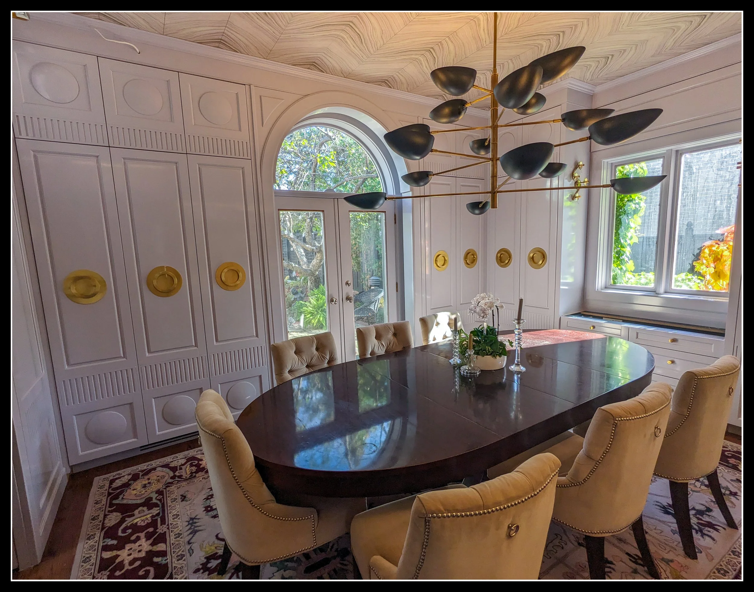 A dining room built from rectangular wall panels, an oval table, and straight architectural lines, softened by round brass pulls, circular raised panels, curved chairs, and an arched doorway that shifts the mood toward warmth and welcome.