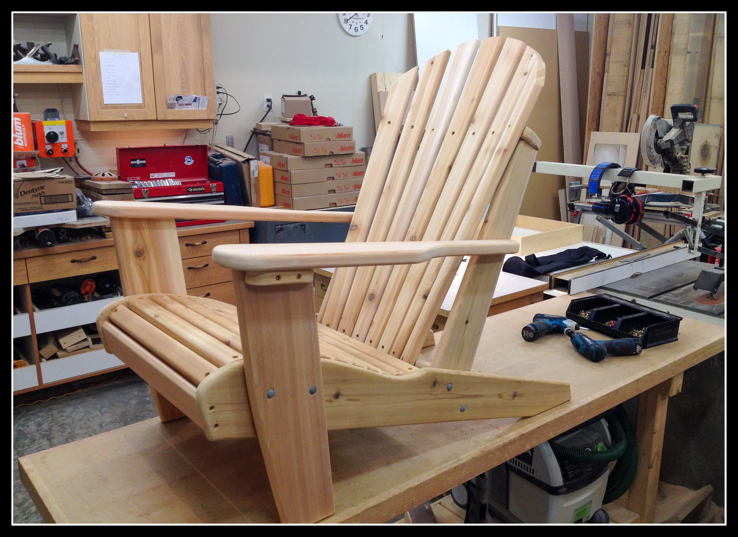 A partially assembled wooden Adirondack chair on a workbench in a woodworking shop with tools and supplies in the background.