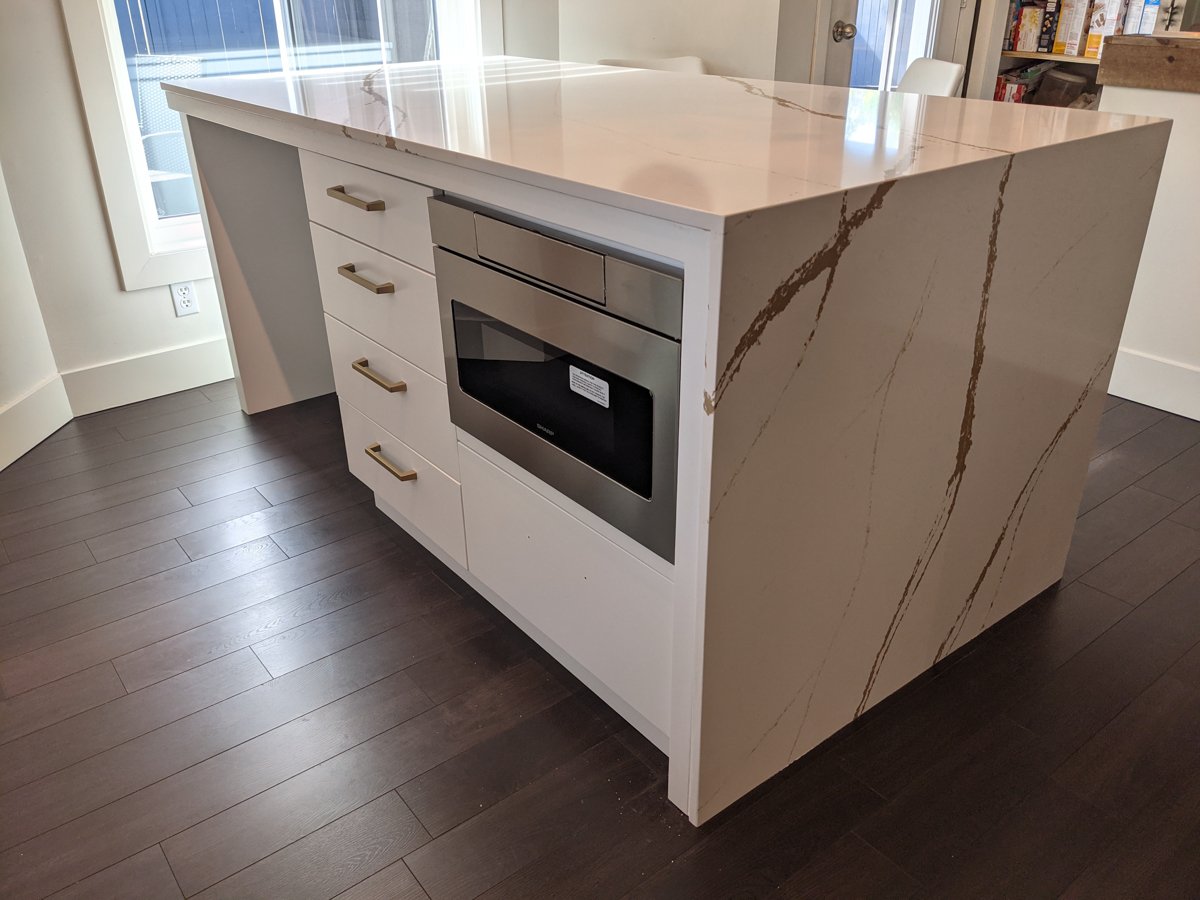 A kitchen island with white drawers, gold handles, and a built-in microwave.