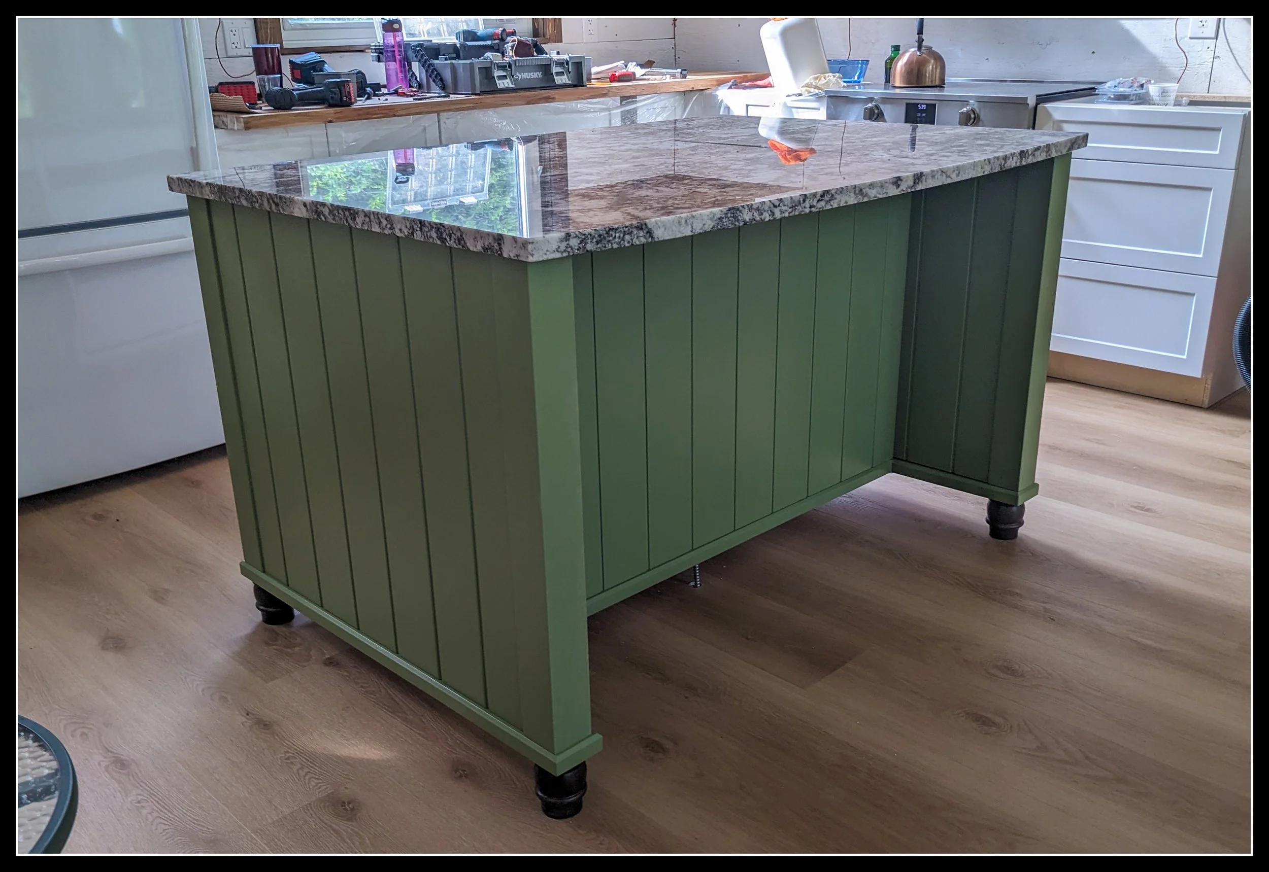 Green bead‑board kitchen island with turned feet and a granite countertop, built in a cottage‑style lake house.