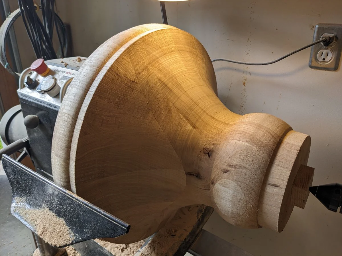 A large wooden table pedestal being turned on a lathe, with wood shavings on the lathe.