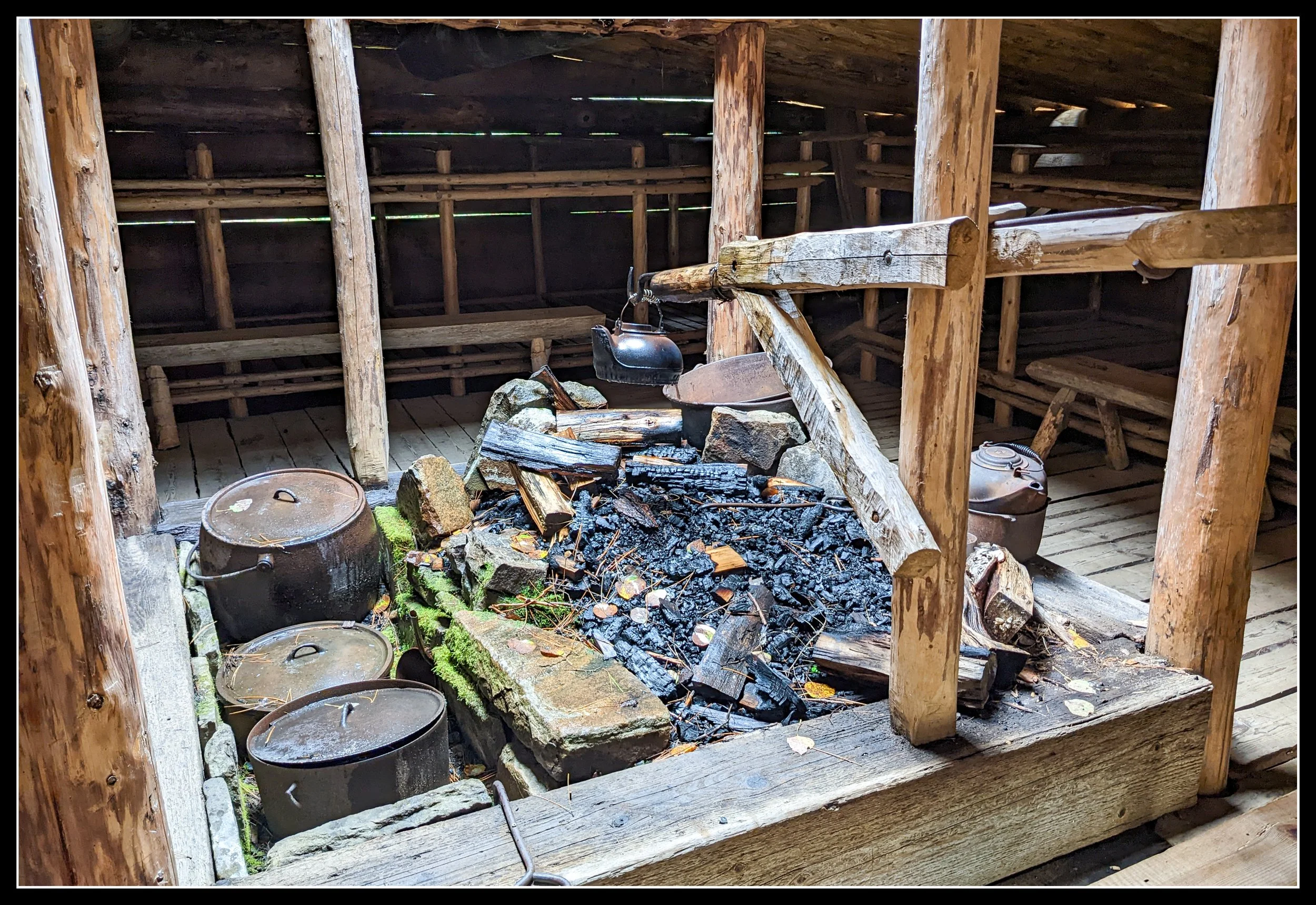 A reconstructed 19th‑century logger’s camp with a central fire pit, rough‑hewn log walls, and cooking kettles, illustrating the type of winter forestry camps used by timber crews supplying York Factory.