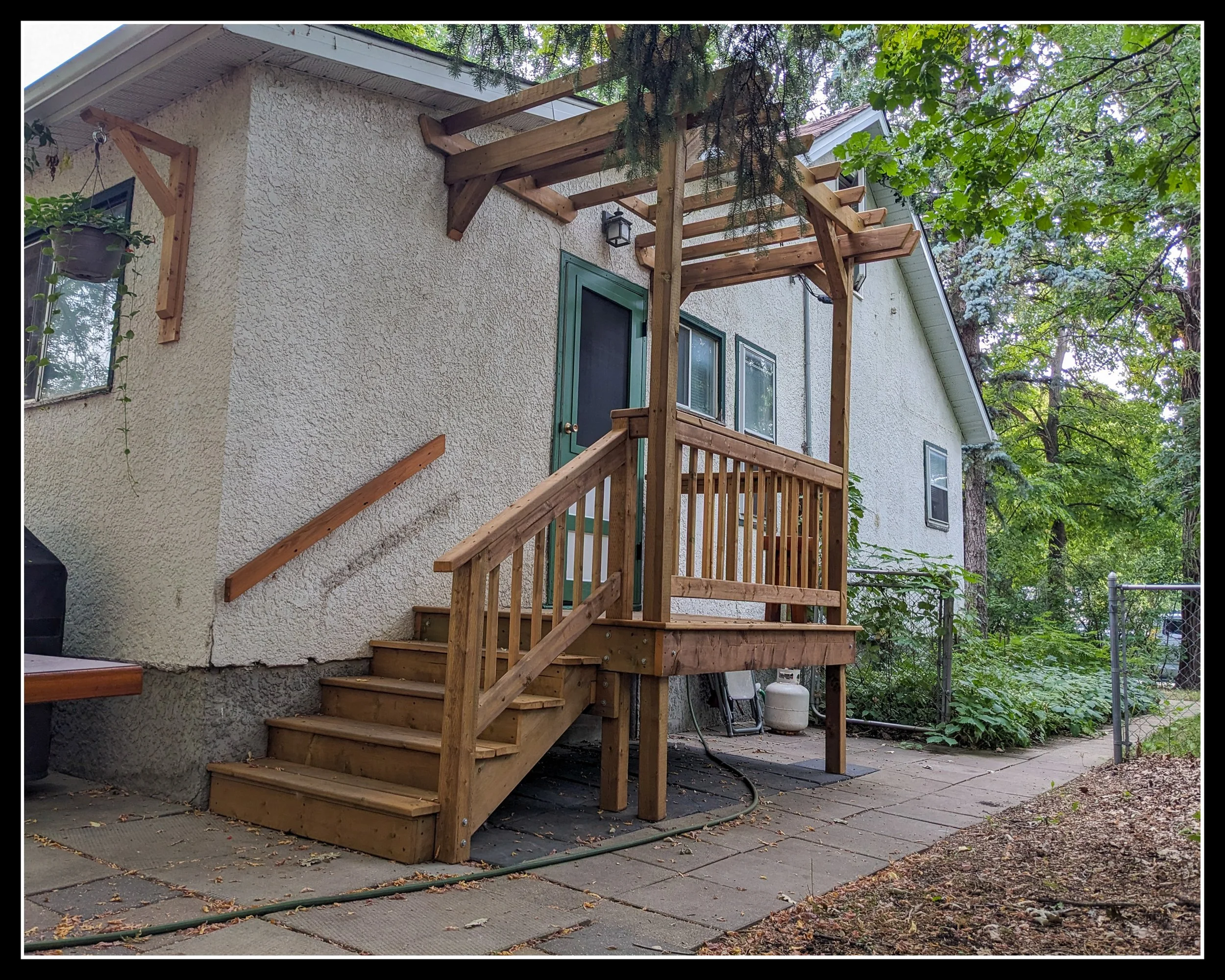 Newly built wooden deck with stairs and a pergola attached to the back of a house, surrounded by trees and greenery.