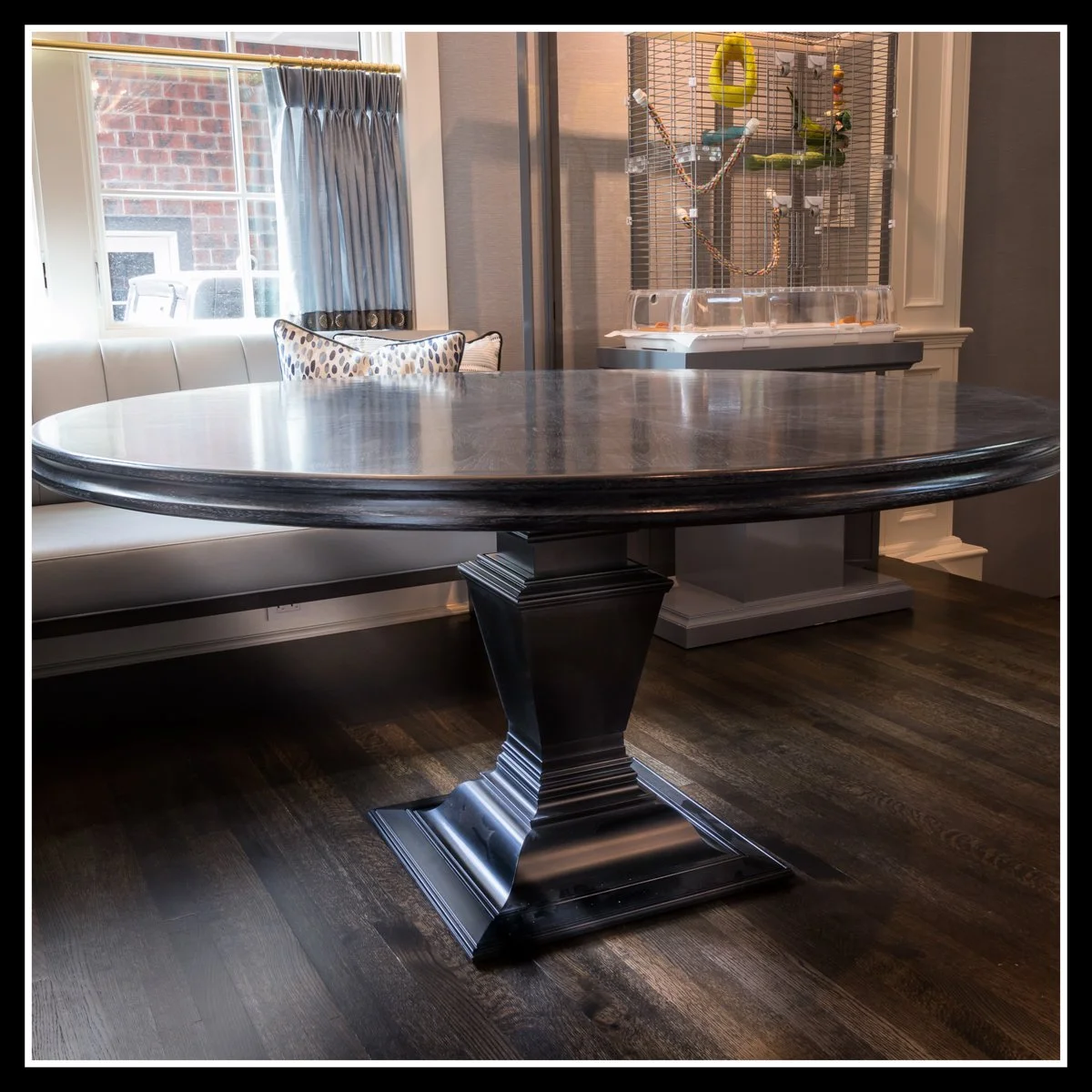 A round walnut wood dining table with a pedestal base, located in a room with a window, curtain, and a computer monitor and a bird cage in the background.