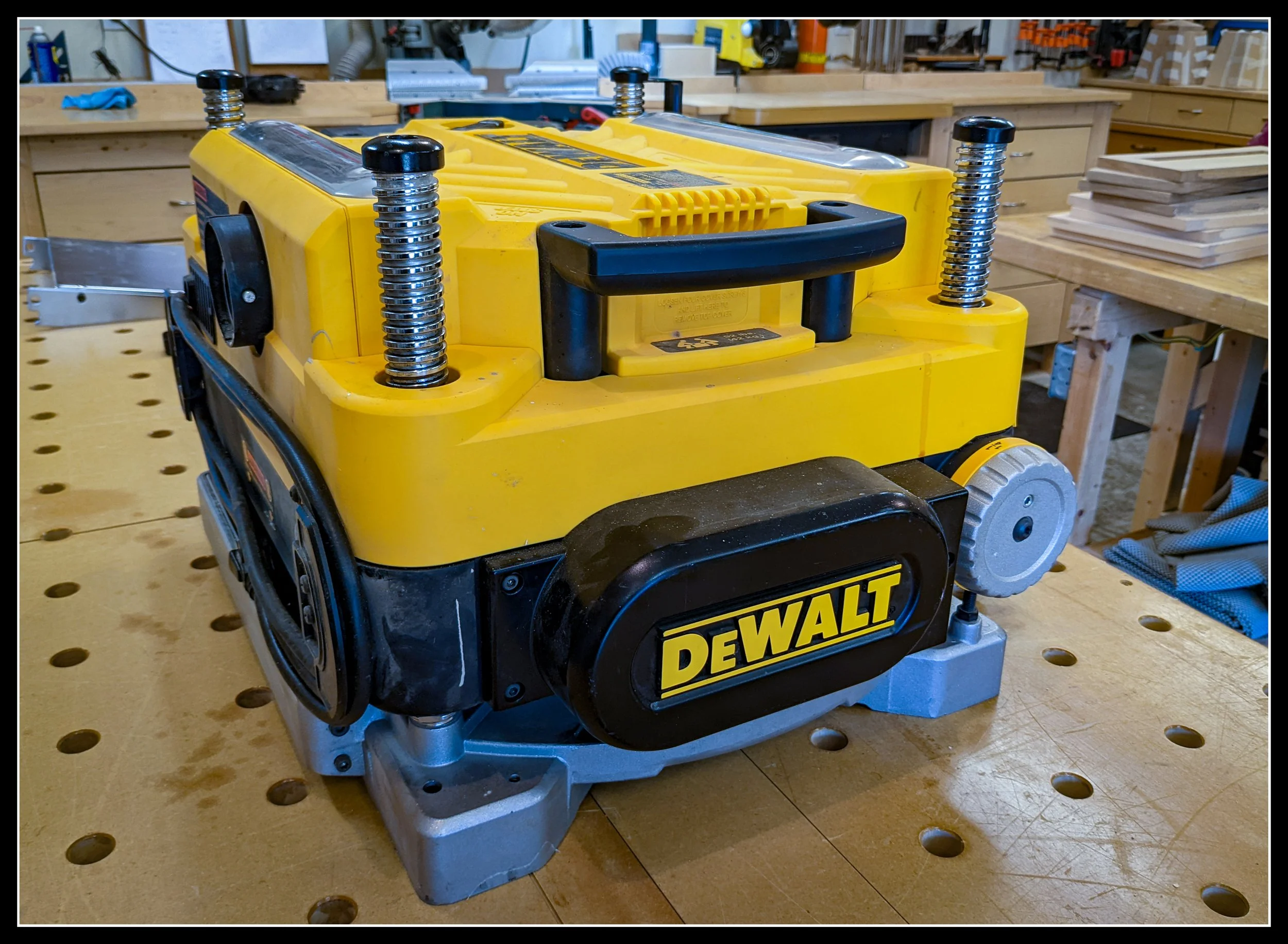 DeWalt 13‑inch portable thickness planer sitting on a perforated workbench in a woodworking shop, showing the height‑adjustment posts, top handle, and yellow housing.